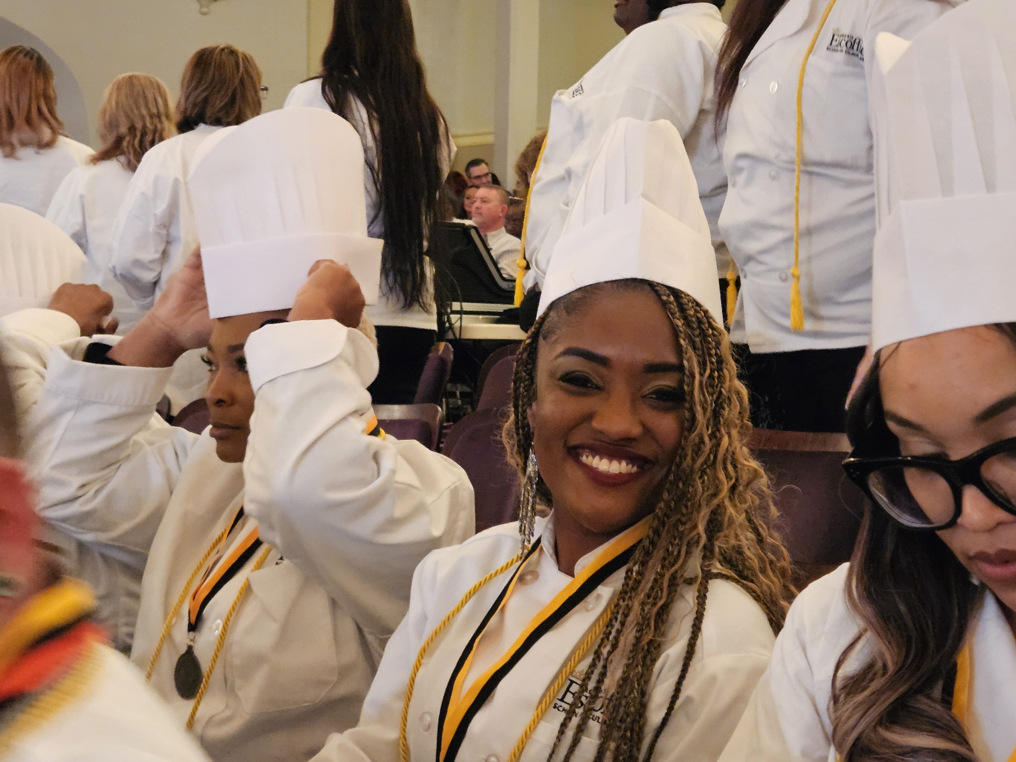 Group of people wearing white chef hats and coats, smiling in a kitchen or culinary classroom.