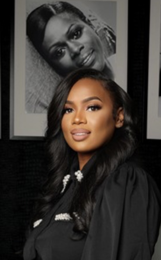 Young woman with long dark hair, smiling, wearing a black outfit, standing in front of black and white photographs.