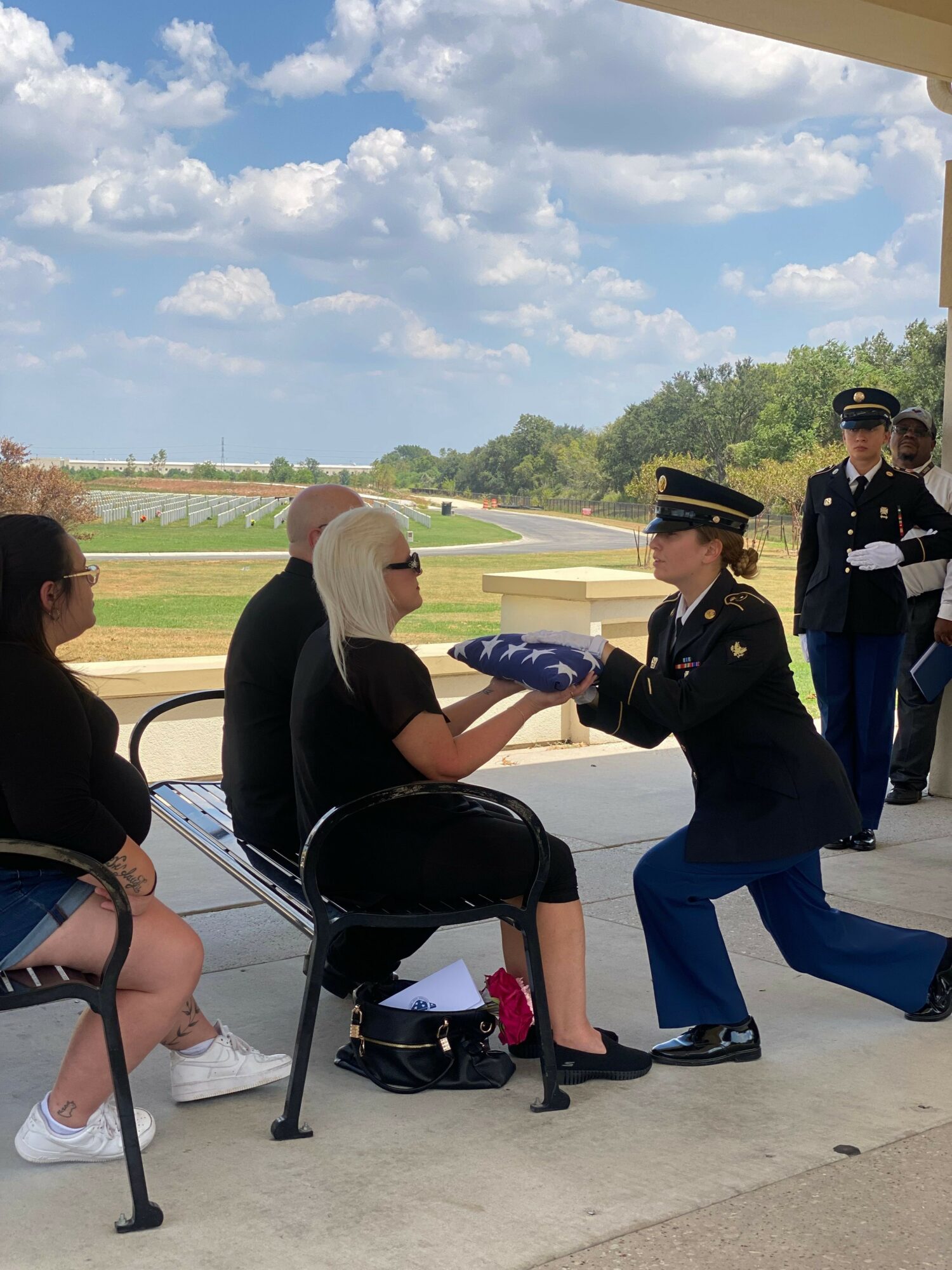 Military personnel presenting an award to a seated woman outdoors with a blue sky and trees in background.