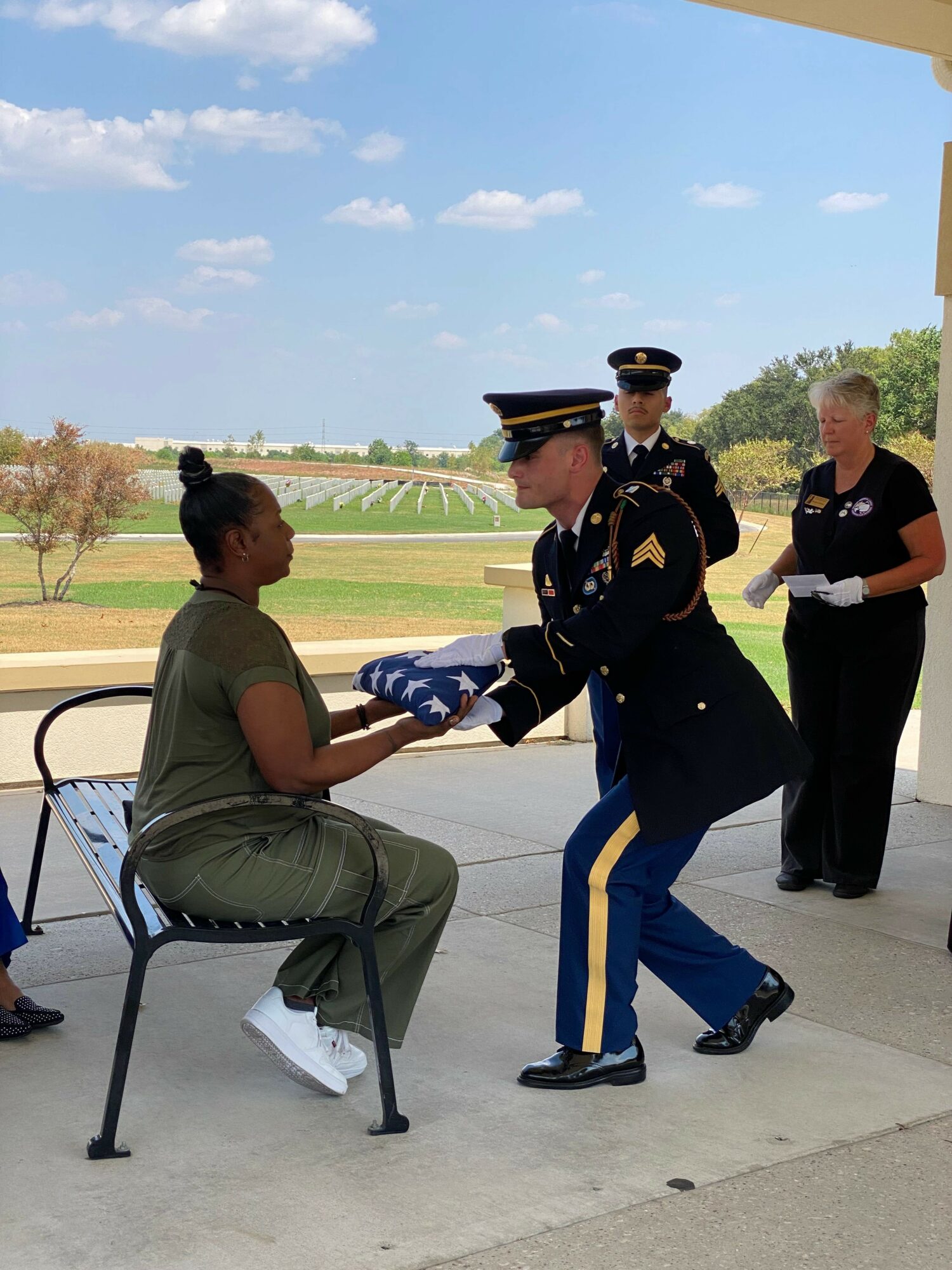 Military personnel presenting an item to a seated woman outdoors, with two people observing nearby and a landscape background.