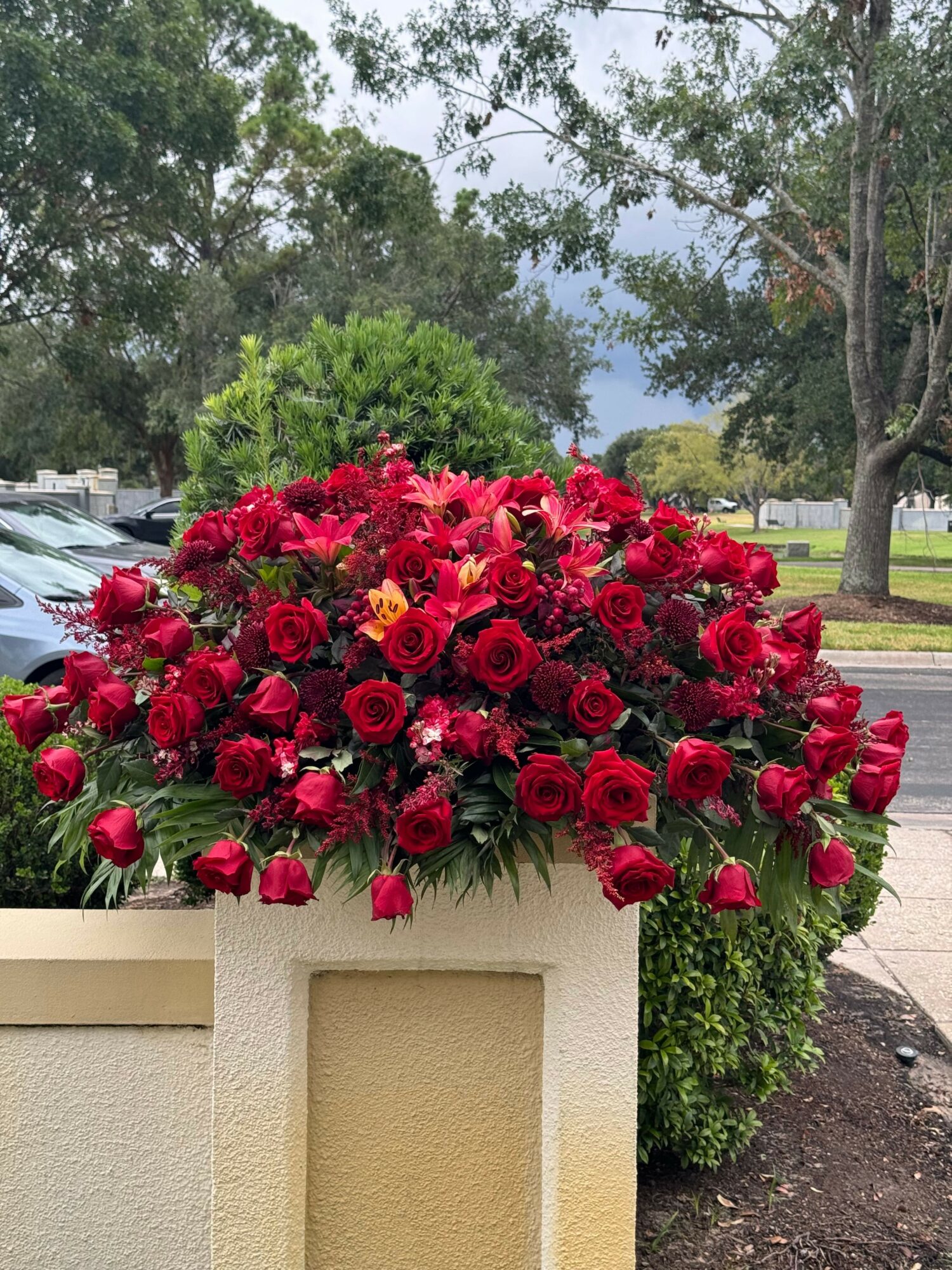 Large bouquet of red roses and flowers on a white planter outdoors, with trees and cloudy sky in background.