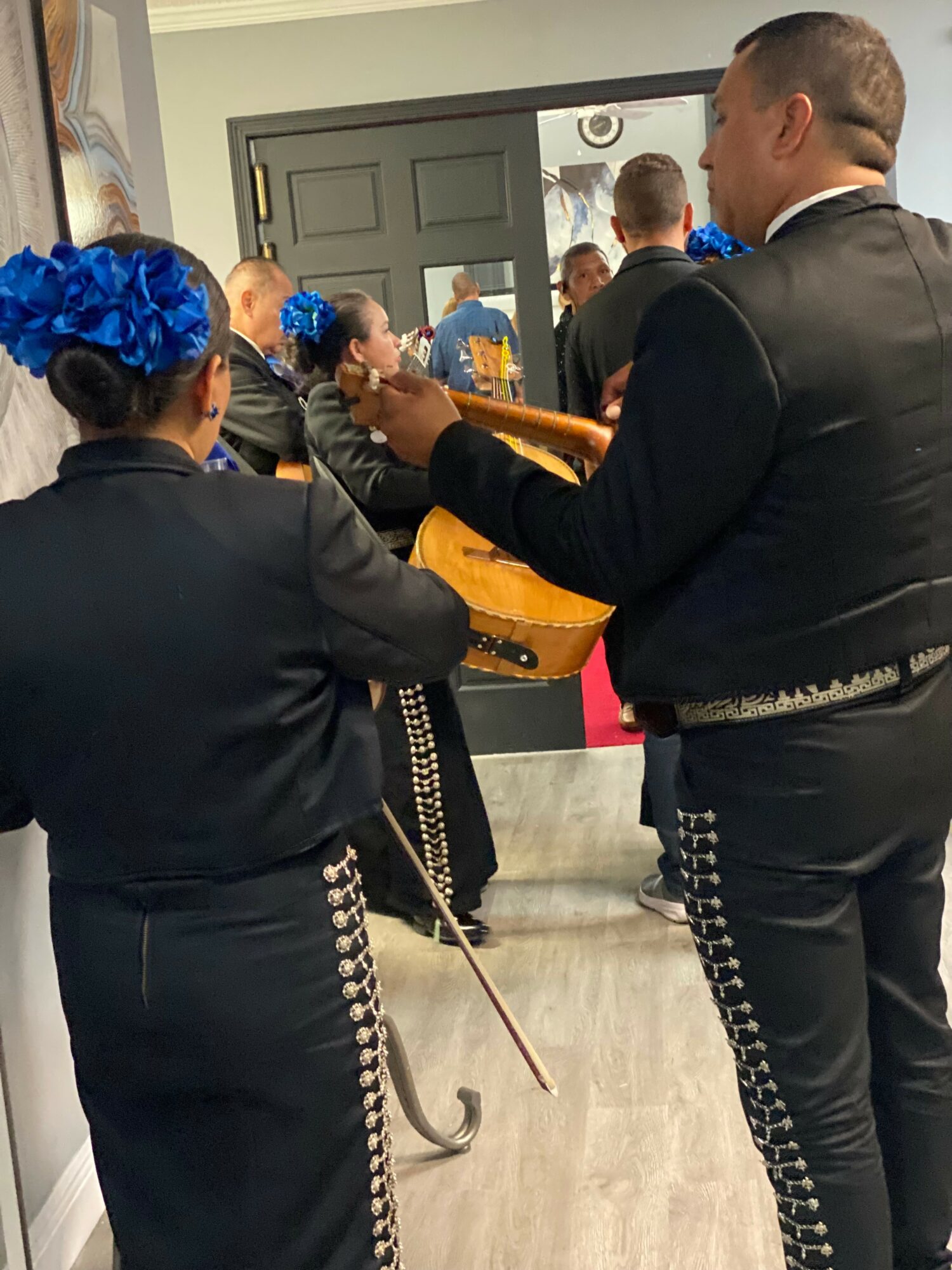 Two mariachi musicians in traditional black outfits with silver accents, one playing a guitar, in a room with a mirror.