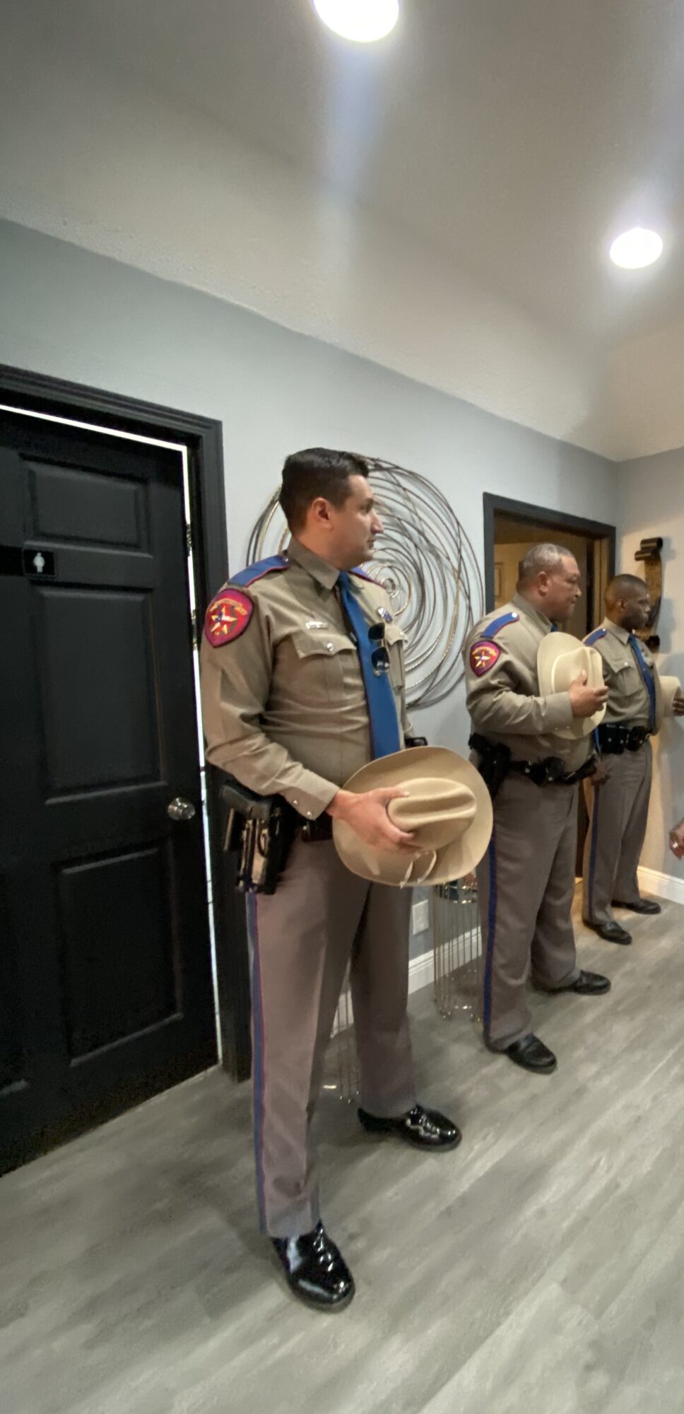 Group of uniformed officers standing indoors, some holding hats, in front of a wall with artwork.