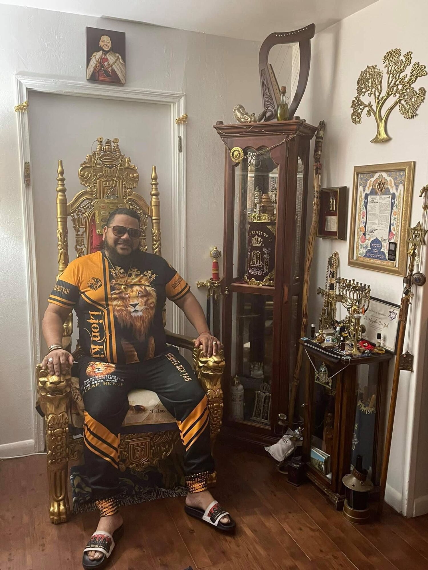 Man with sunglasses sitting on ornate golden throne in a room with various decorative items and framed pictures.
