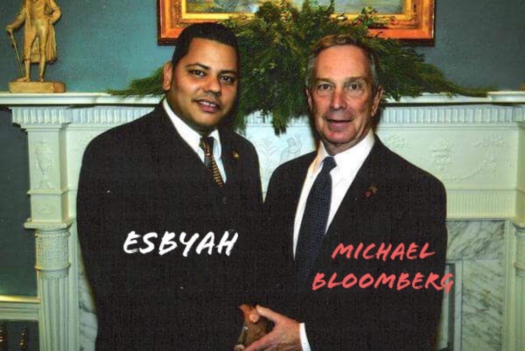 Two men in suits shaking hands in front of a fireplace with a painting and greenery above.