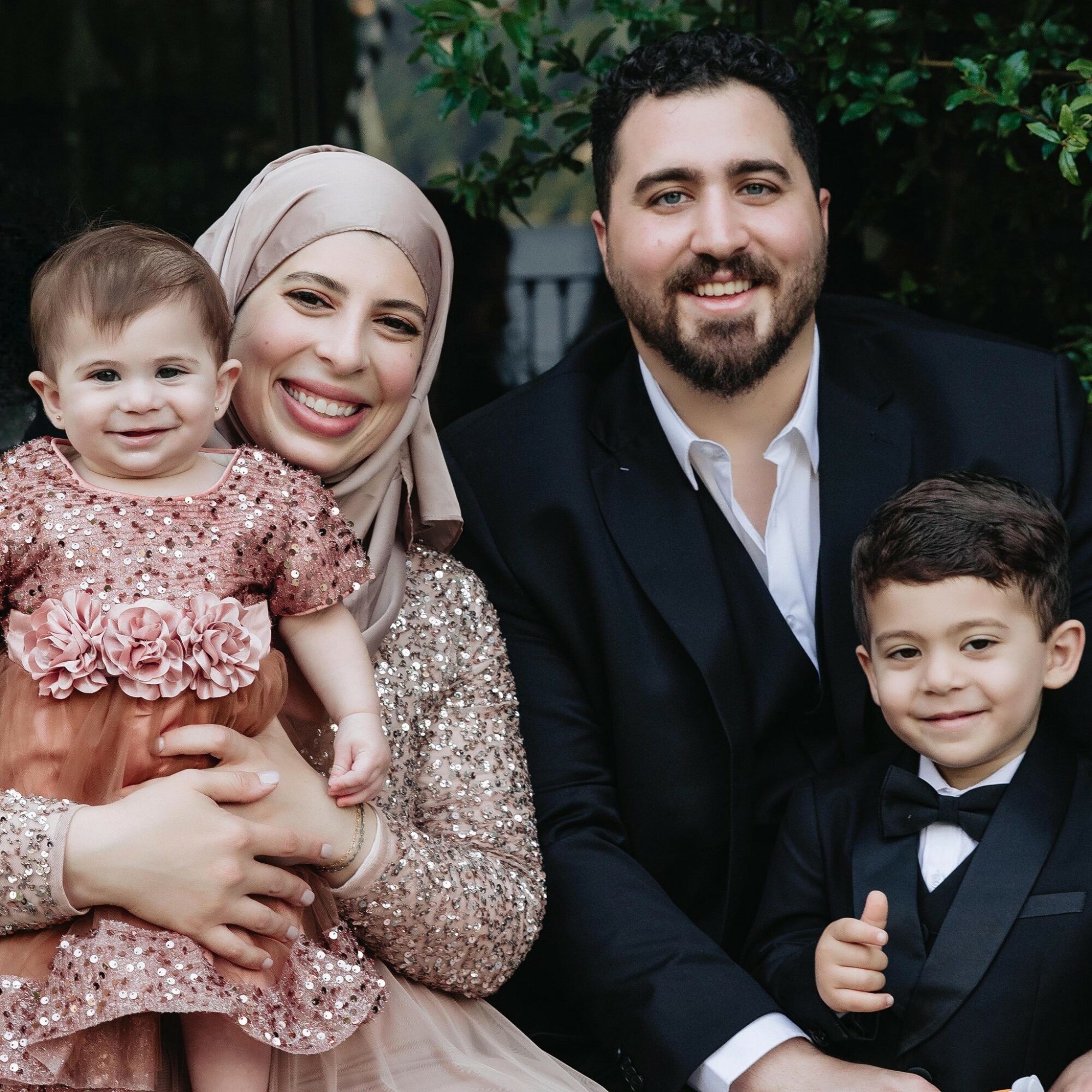 Family of four smiling outdoors, woman in hijab, man in suit, two children, greenery background.