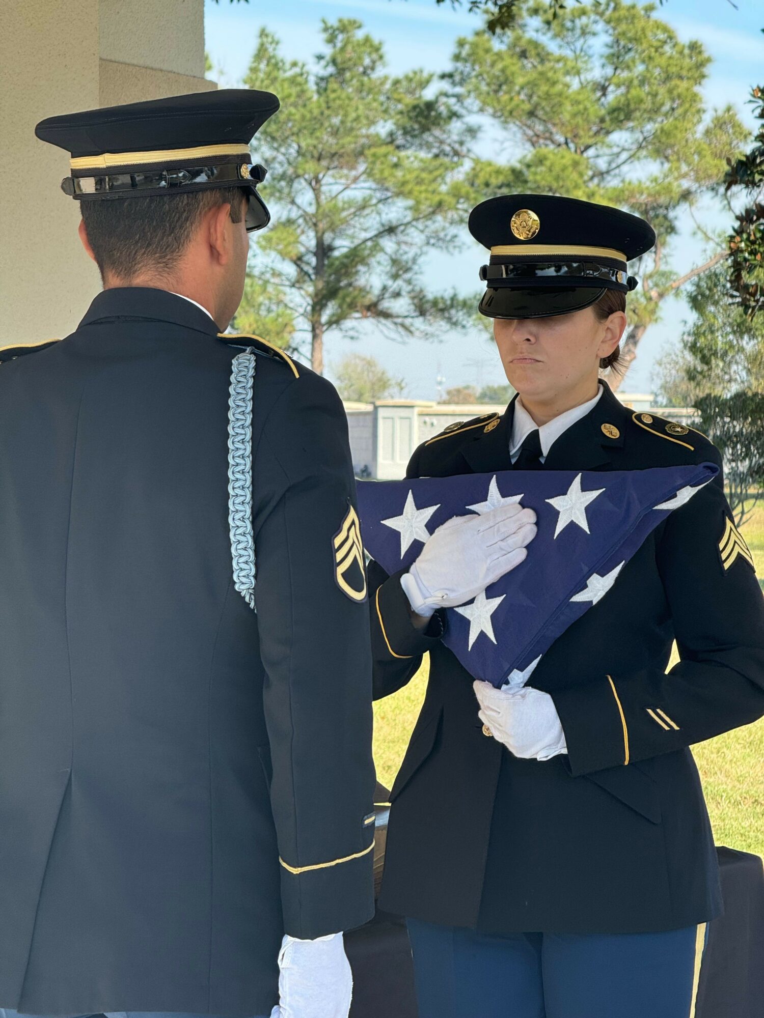 Two uniformed officers holding a folded American flag outdoors with trees in the background.