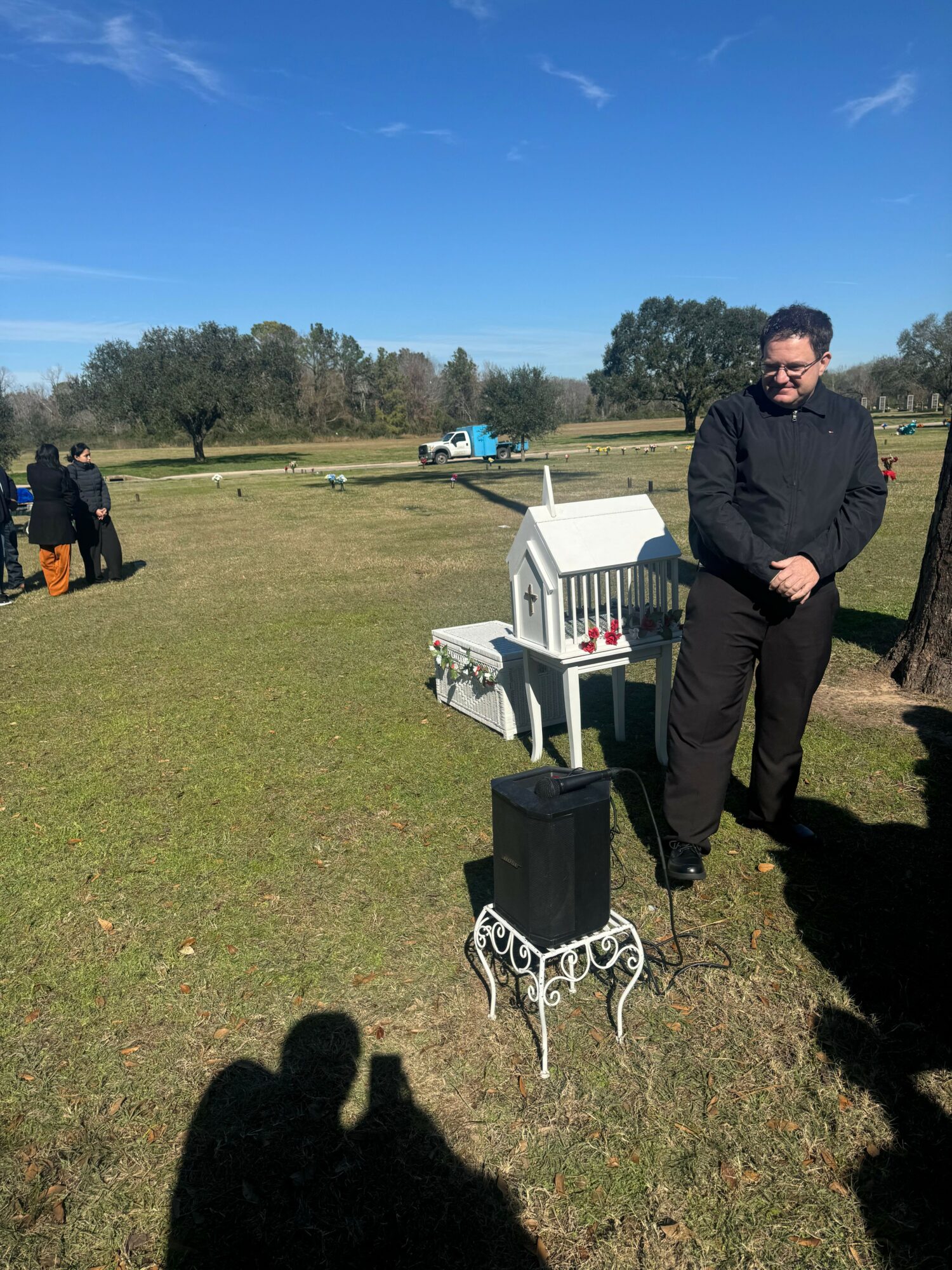 Man in black jacket standing outdoors on grassy field with trees and blue sky in background.