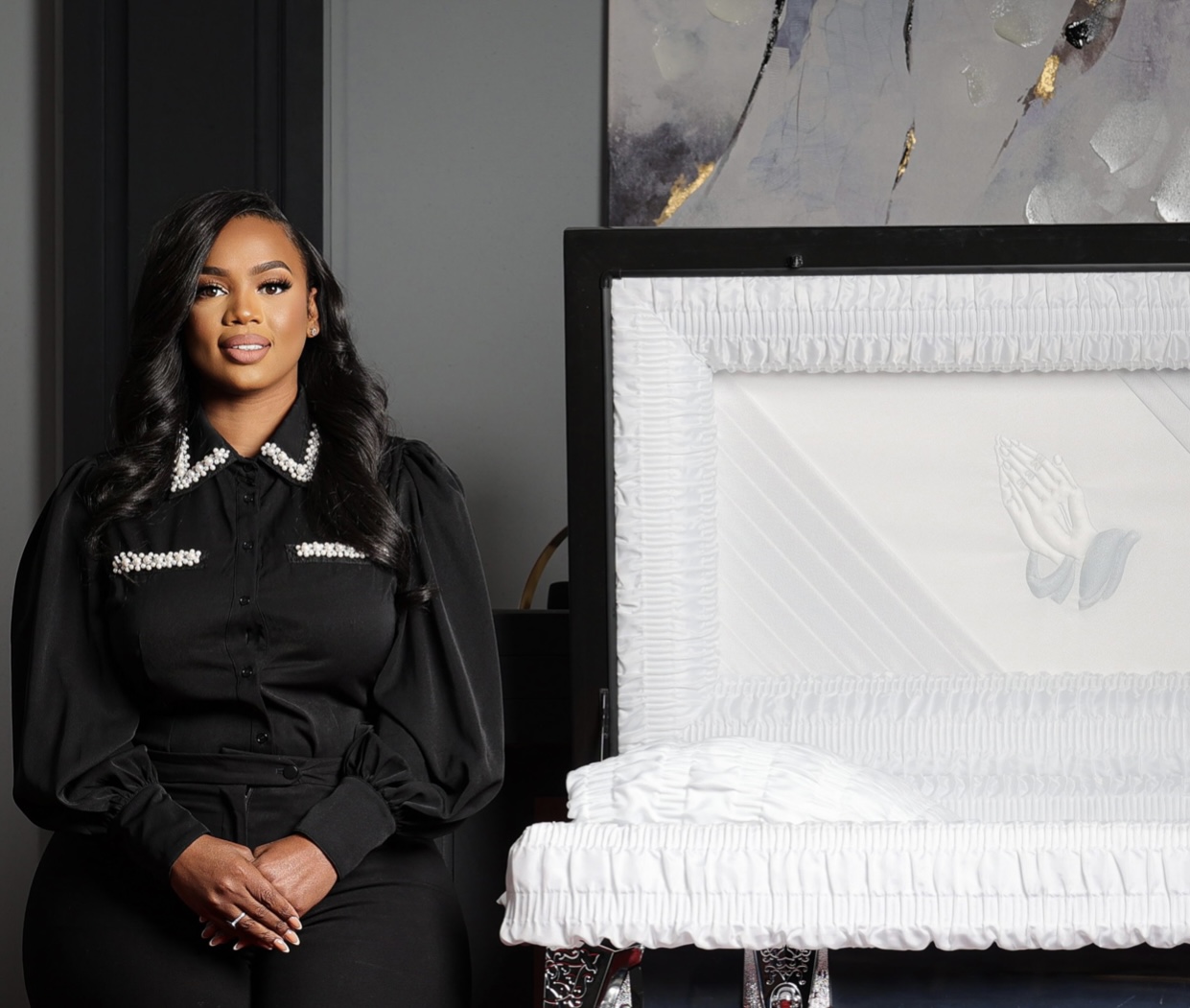 Woman standing next to a white casket with a black frame, in a room with abstract wall art.