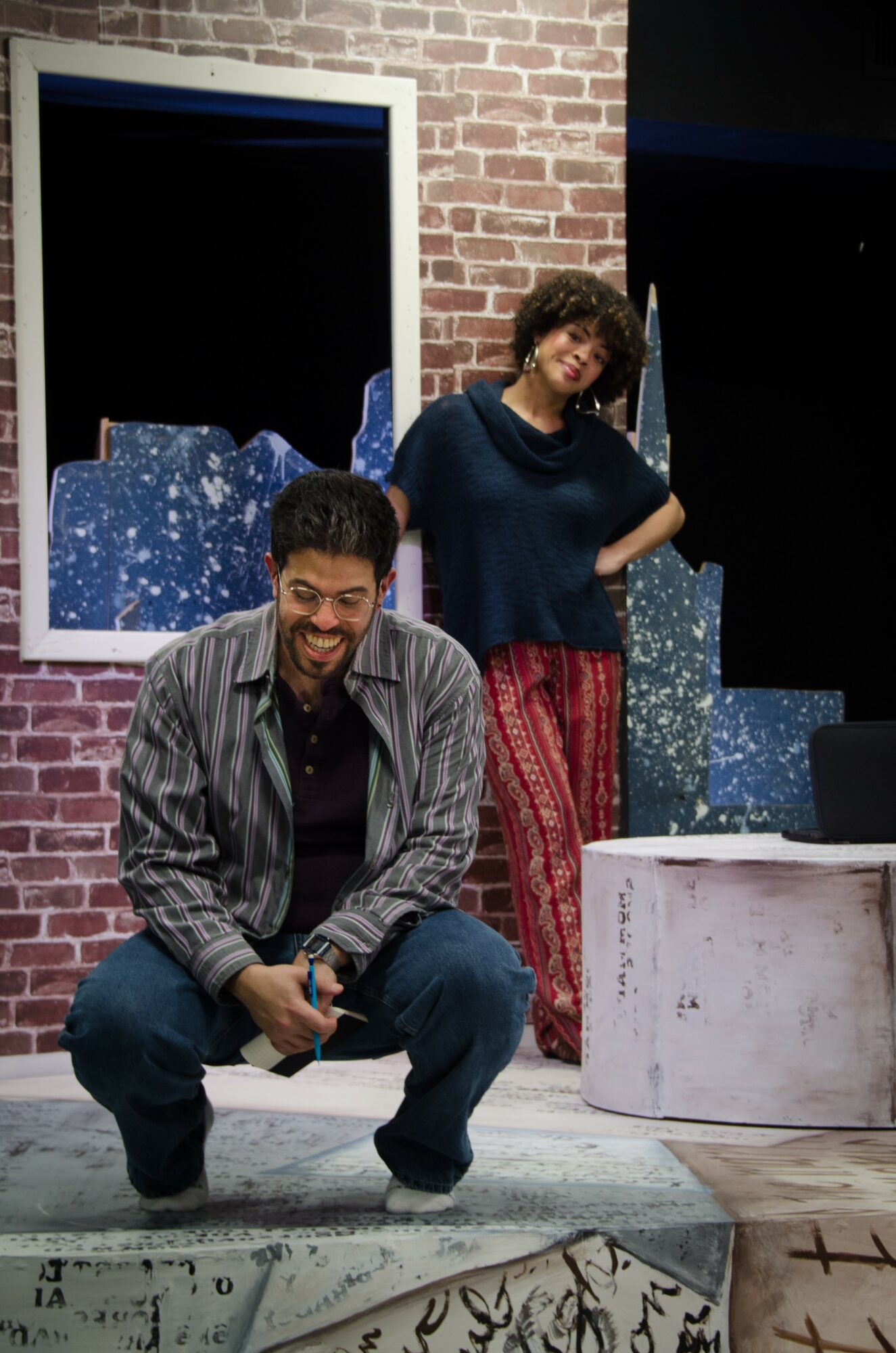 Two people, a man crouching and a woman standing, in front of a brick wall with window and decorative objects.