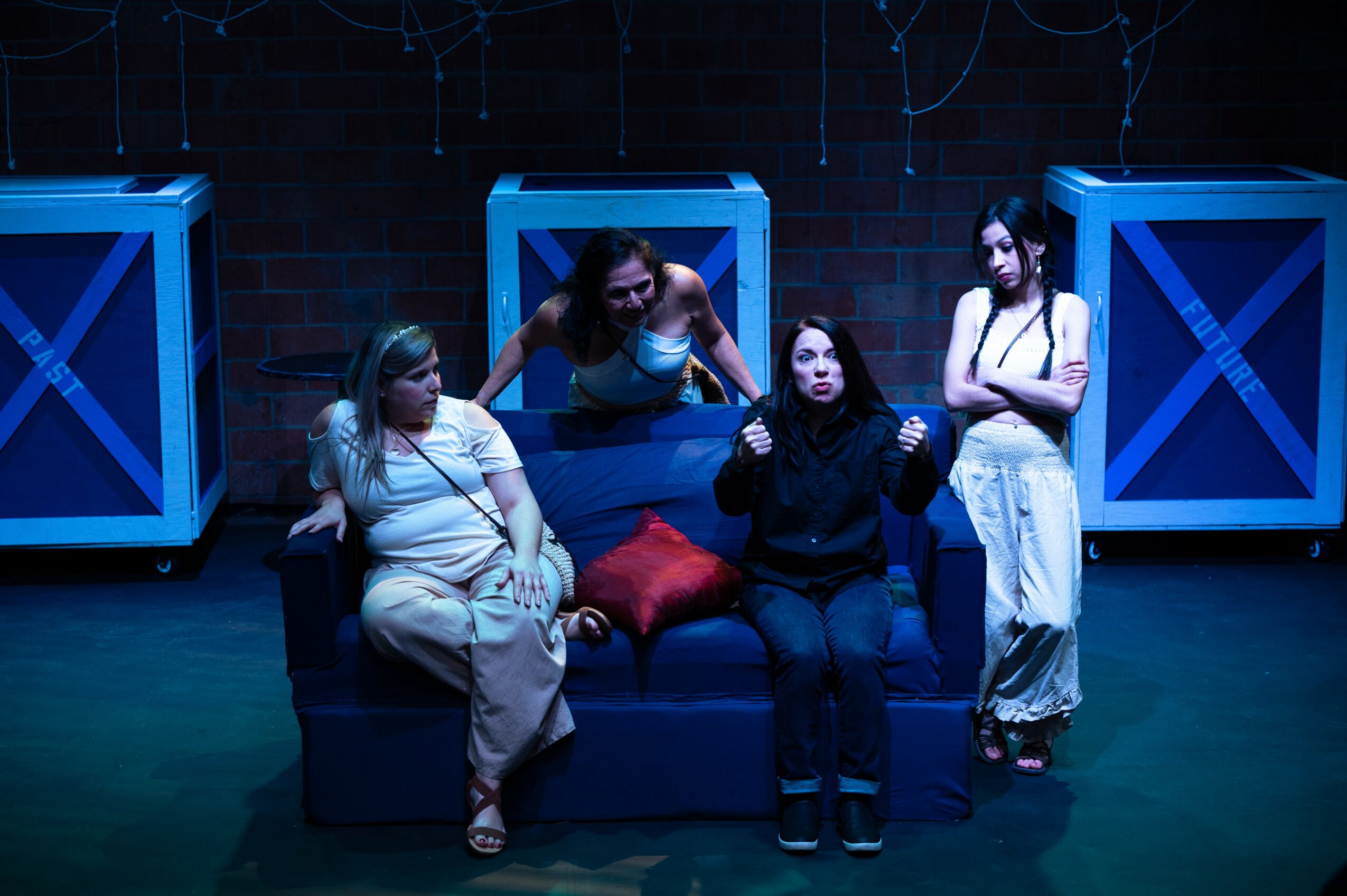Four women on a stage with blue crates in the background, one sitting on a sofa, three standing around her.