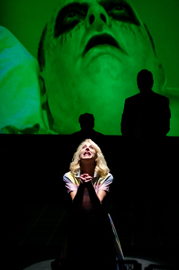 A woman with blonde hair and a colorful shirt looks up with hands clasped, in front of a large screen showing a face.