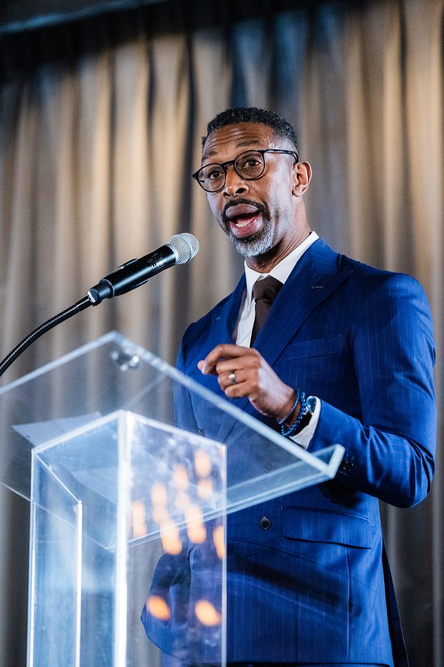 Man in a blue suit speaking at a podium with a microphone, curtains in the background.