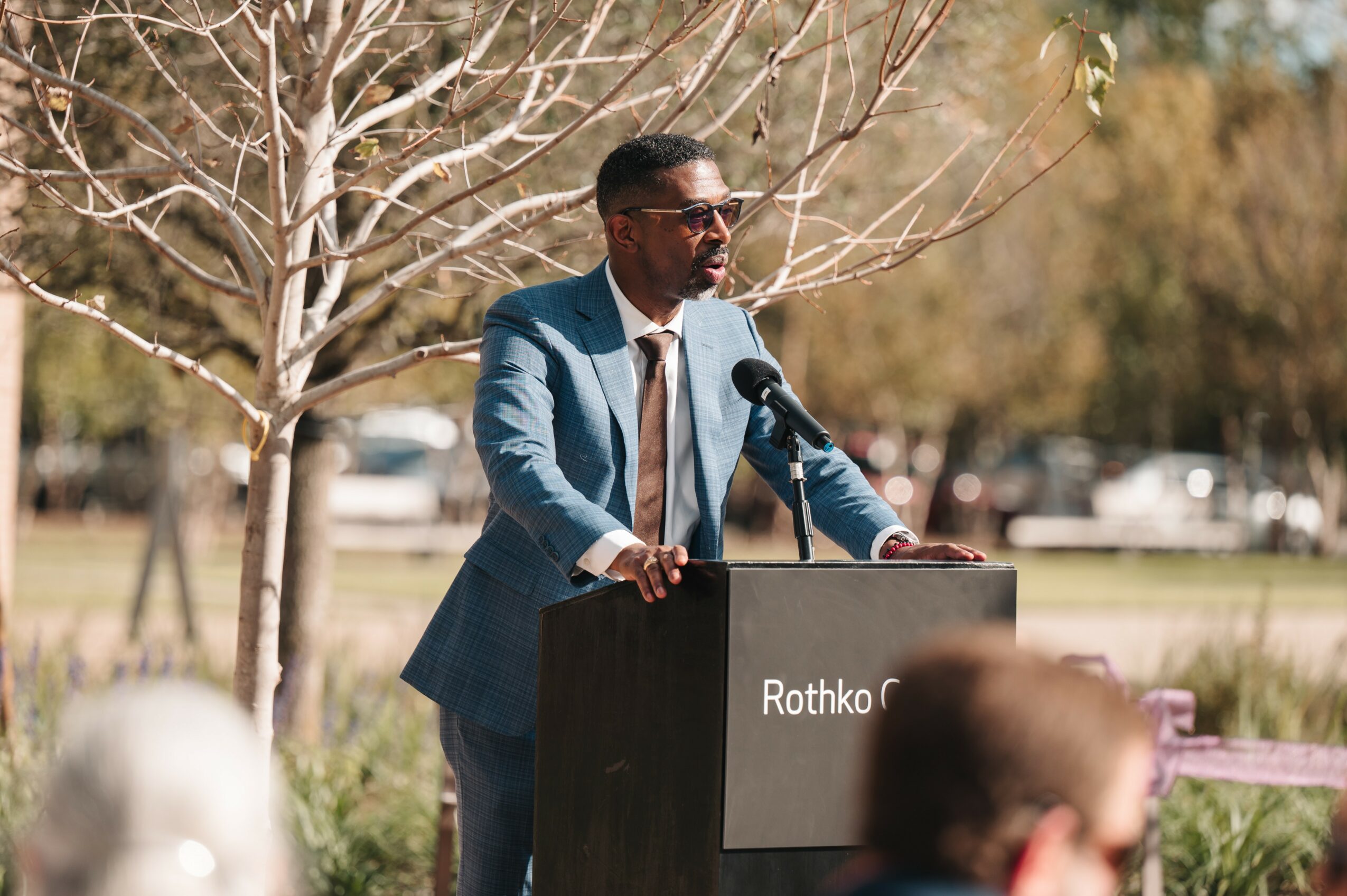 Man in suit speaking at podium outdoors with leafless tree behind him.