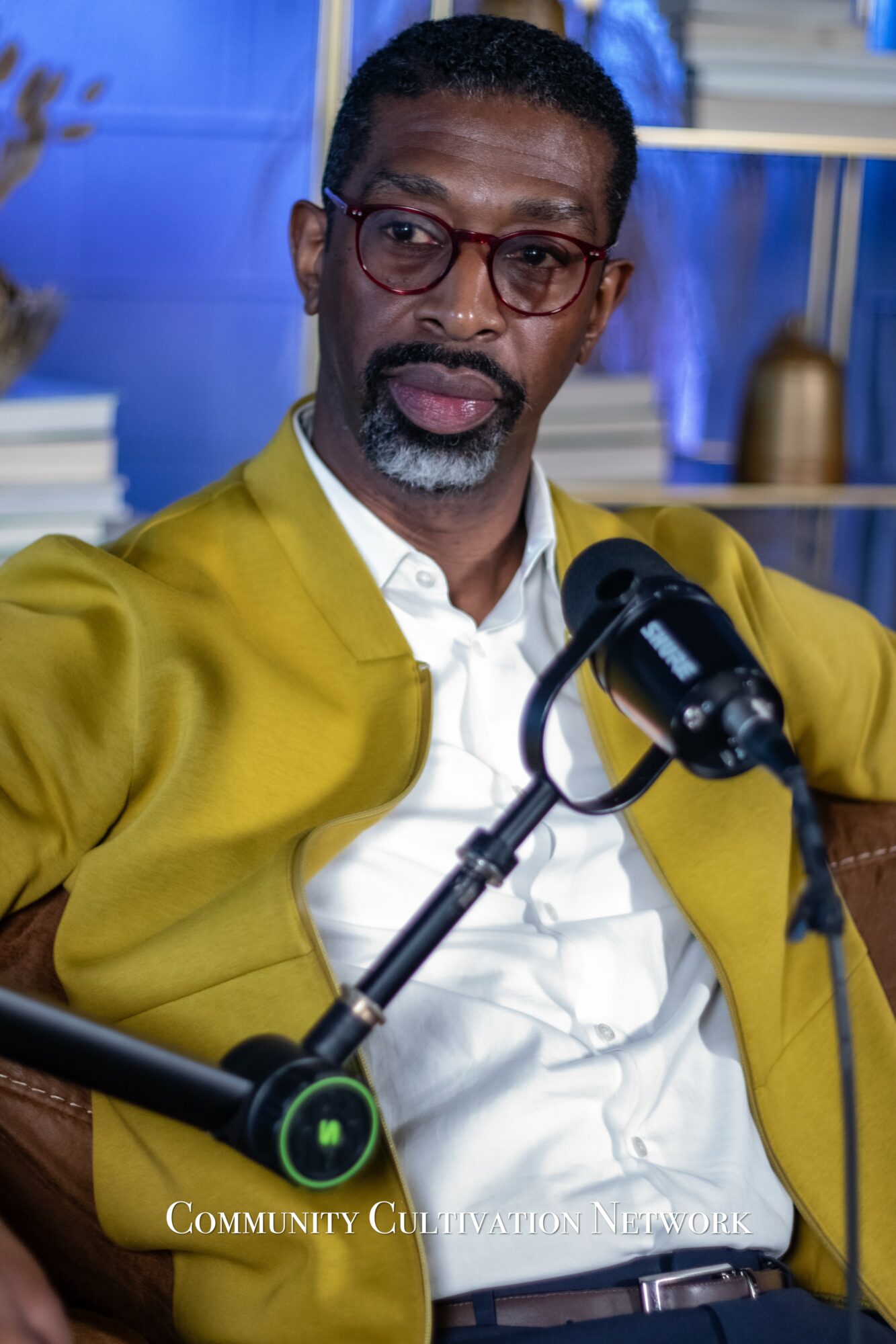 Man with glasses and beard wearing a yellow jacket and white shirt, sitting at a table with a microphone.