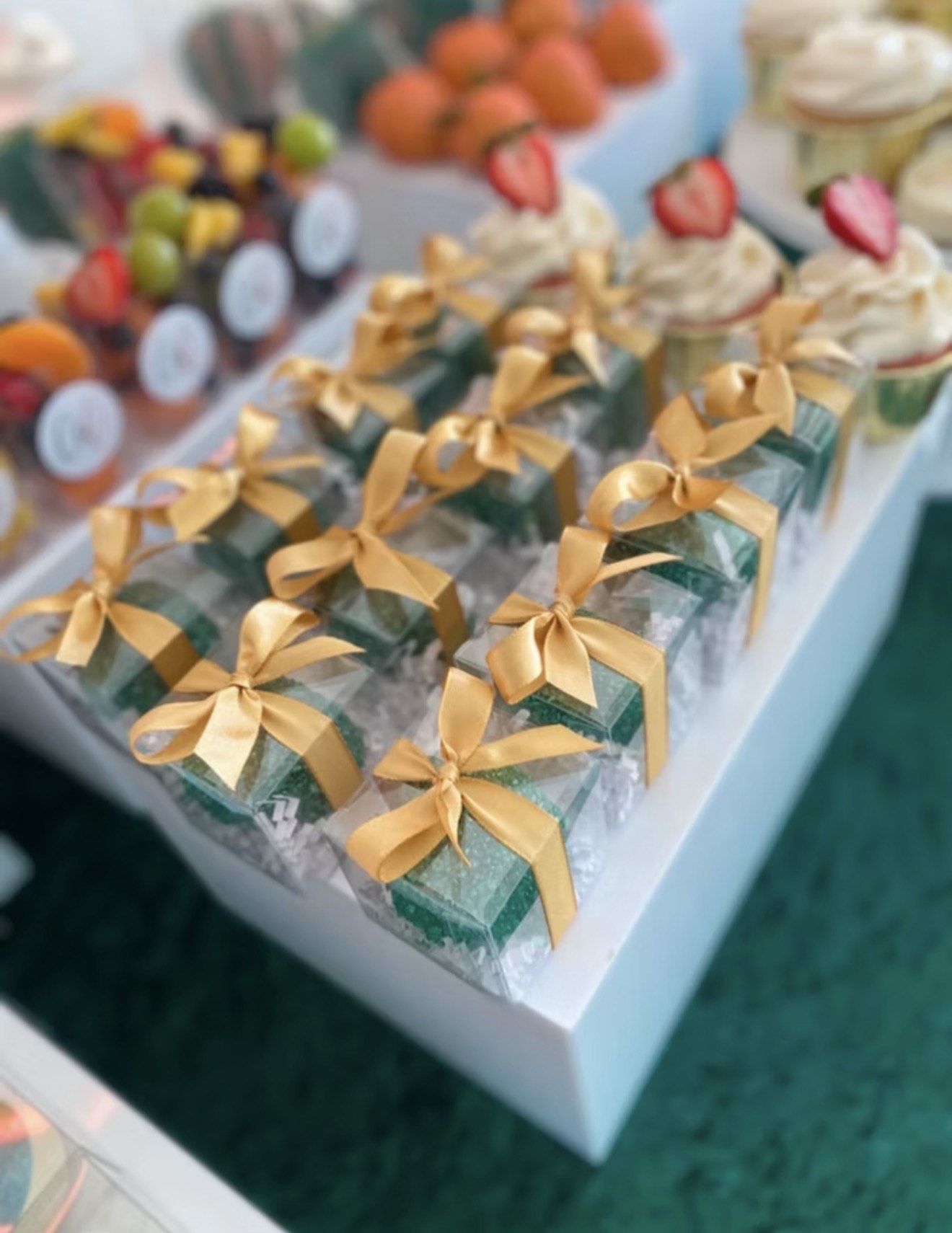 Small gift boxes with gold ribbons arranged on a white table, with cupcakes and other treats in the background.