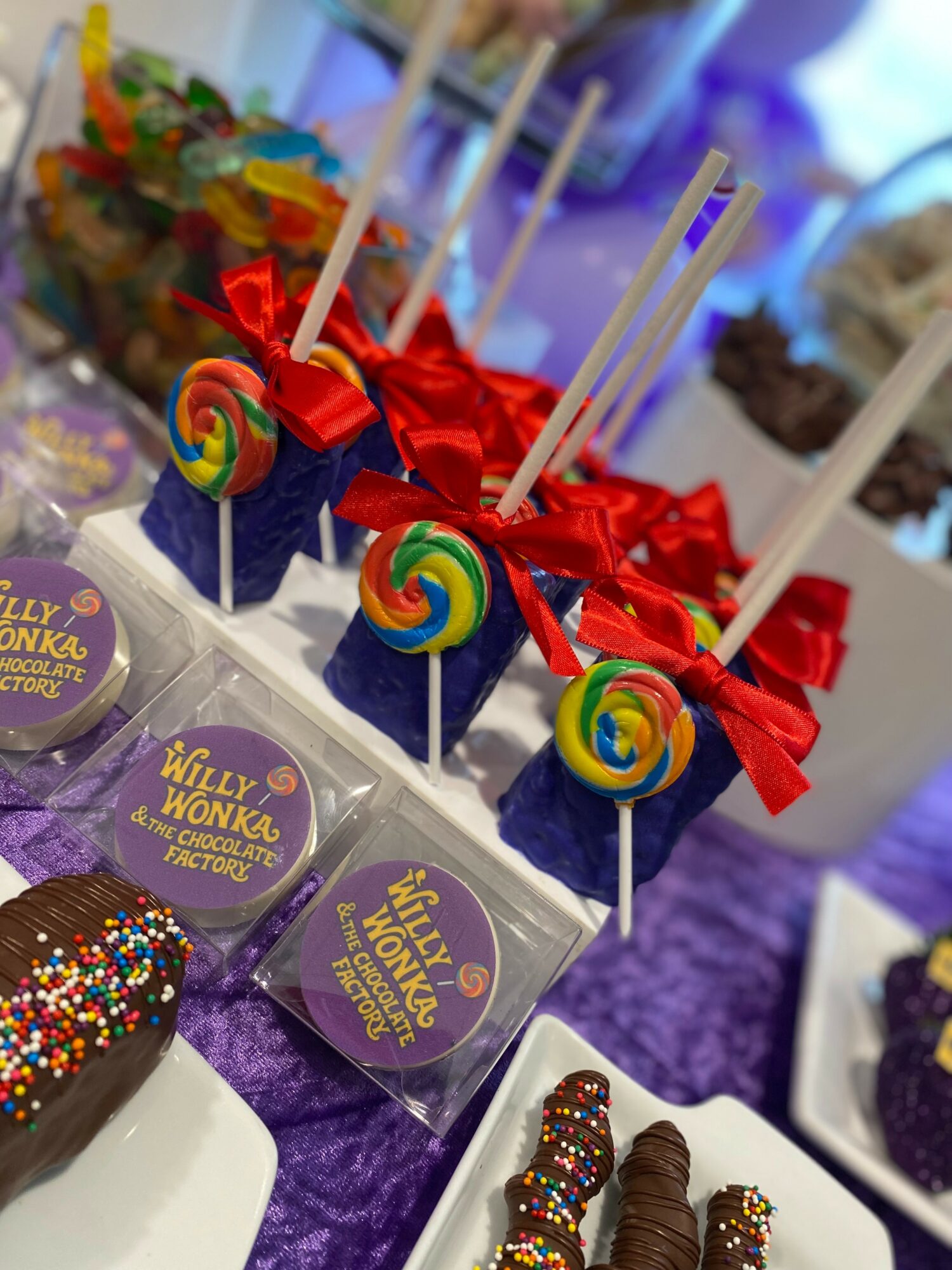 Decorated cake pops with red ribbons and colorful lollipops on sticks, displayed on a table with purple tablecloth.