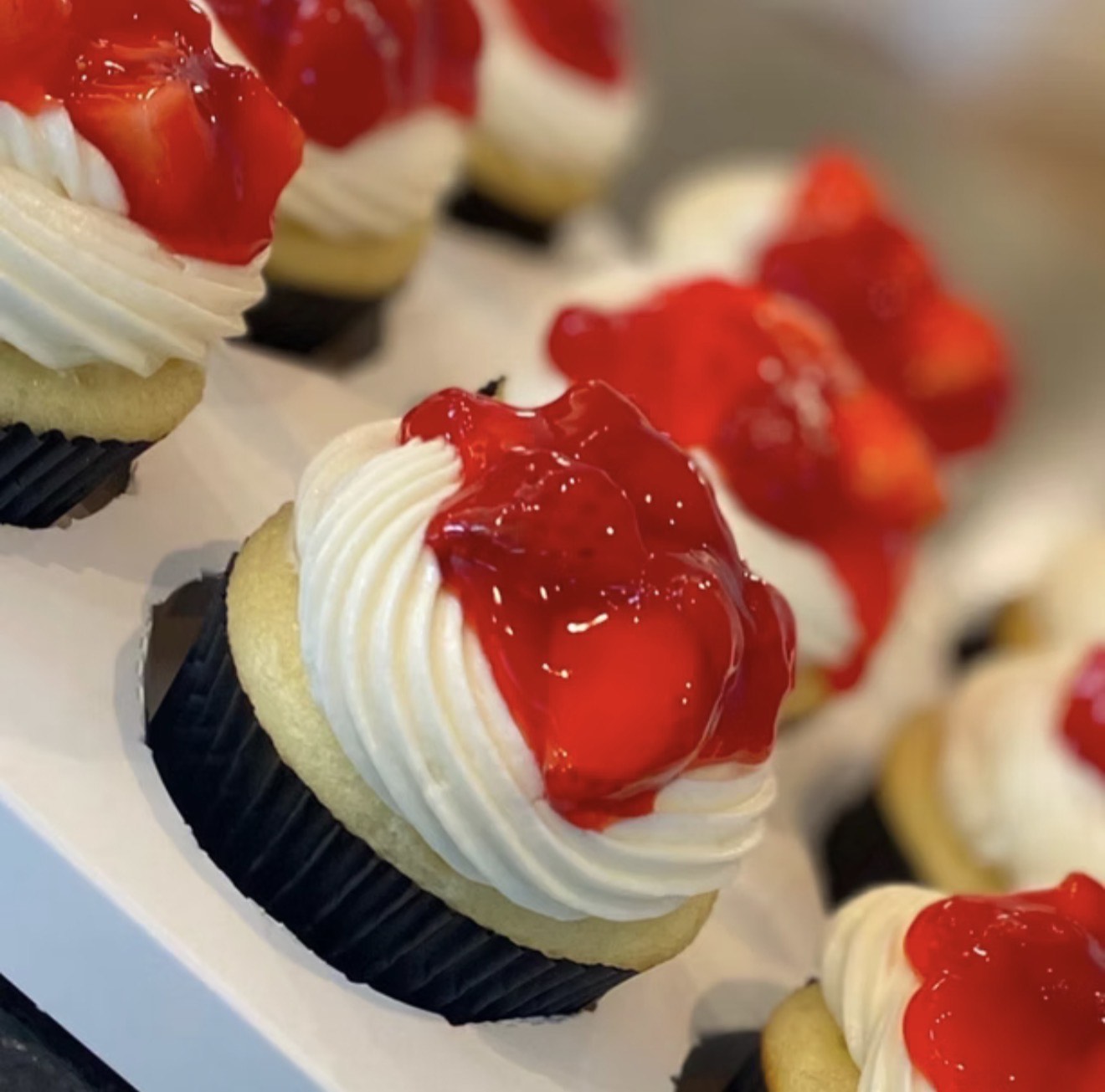 Cupcake with white frosting and red fruit topping, in black paper liner, on white surface.