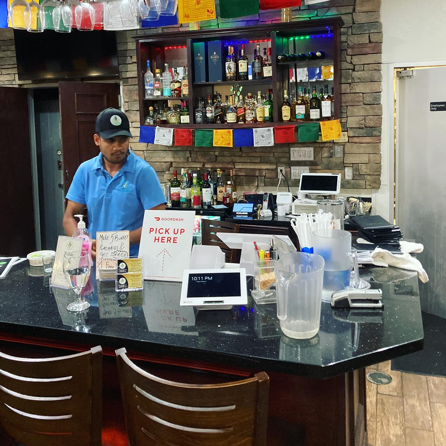 Barista in blue shirt and cap behind counter with bottles and equipment, bar area with colorful decorations, and chairs in foreground.