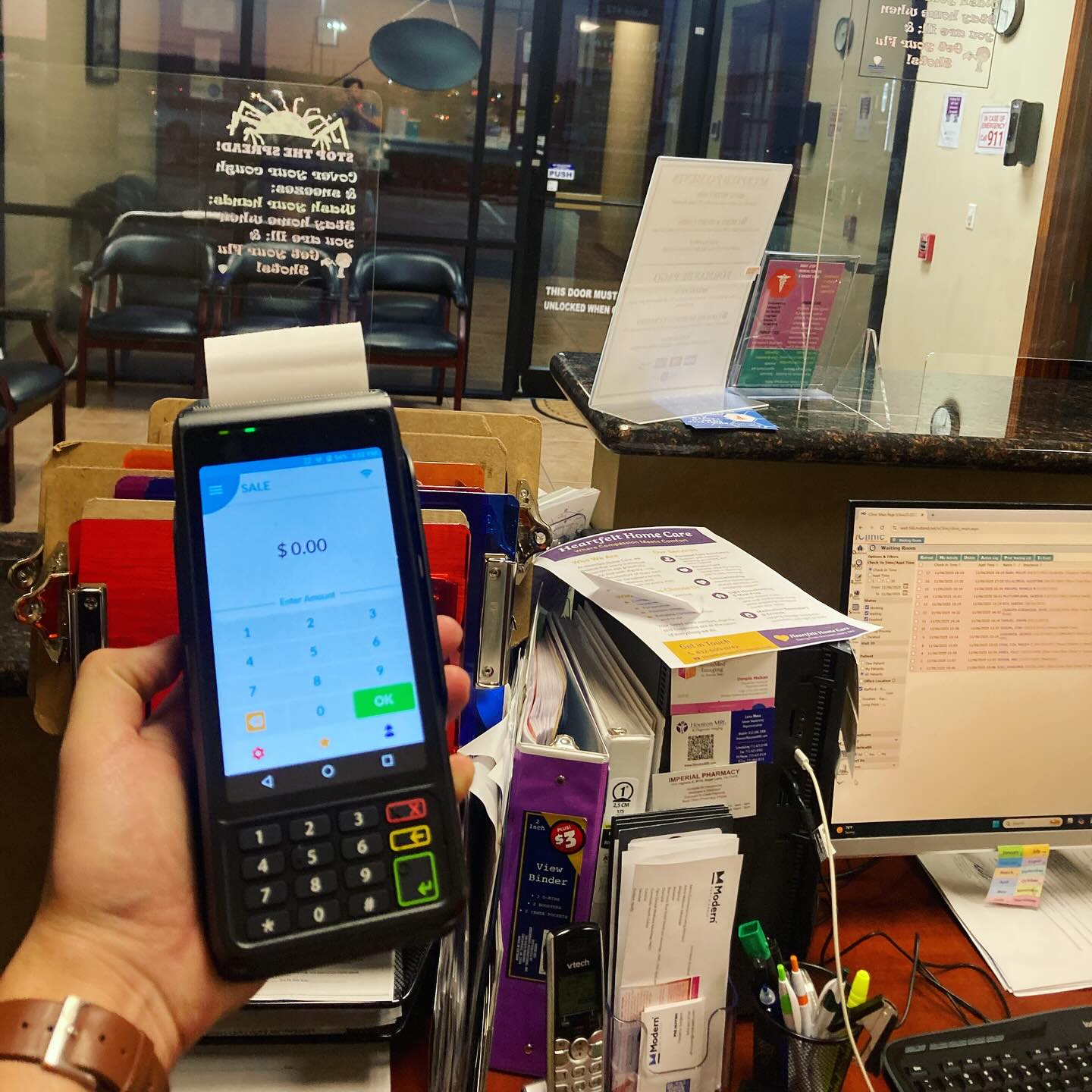 Hand holding a smartphone displaying a payment app, with a reception desk and office in the background.