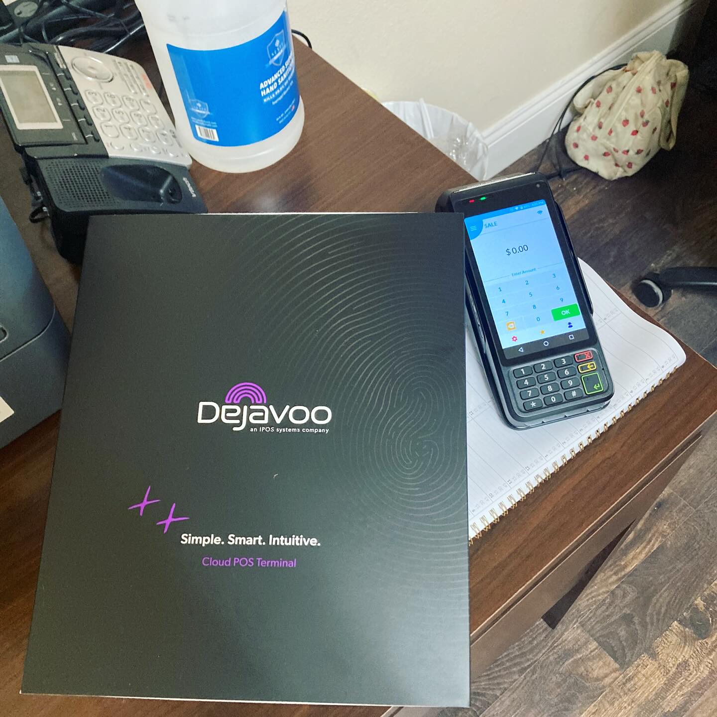 Dejavoo payment terminal box on a wooden desk with a smartphone, notebook, and a bottle in the background.