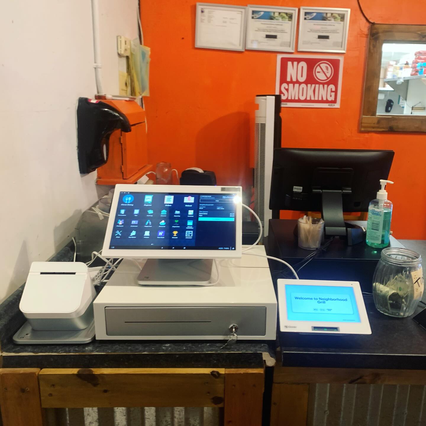 Cash register with touchscreen monitor, card reader, and receipt printer on a wooden counter, with a no smoking sign and sanitizer nearby.