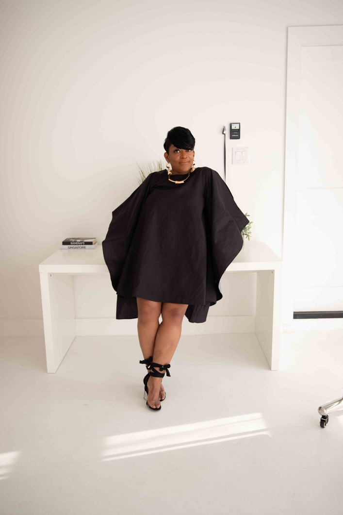 Person standing in front of a white wall, wearing a black oversized coat, black hat, and high heels.