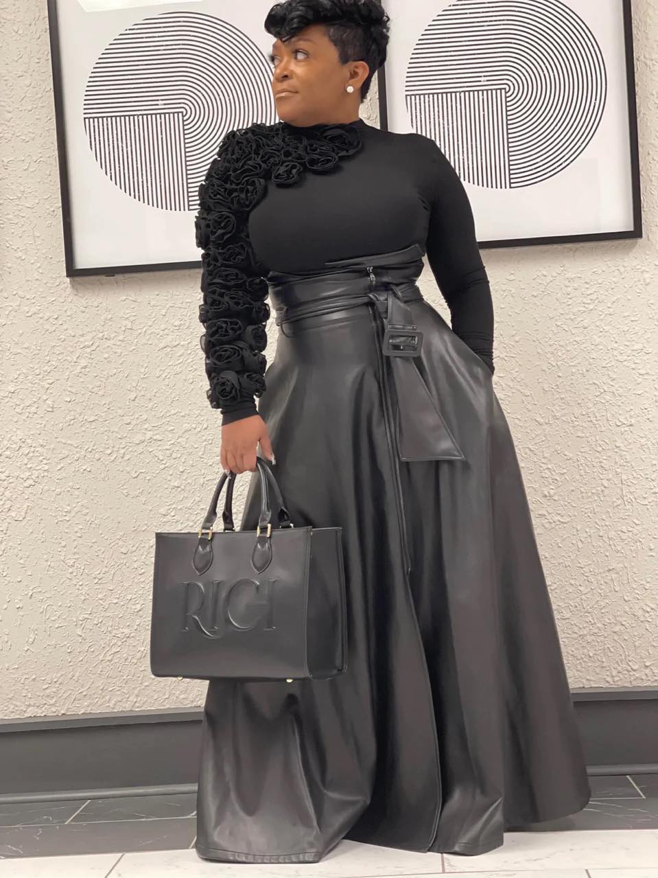 Woman in black outfit holding a large black bag, standing indoors in front of abstract wall art.