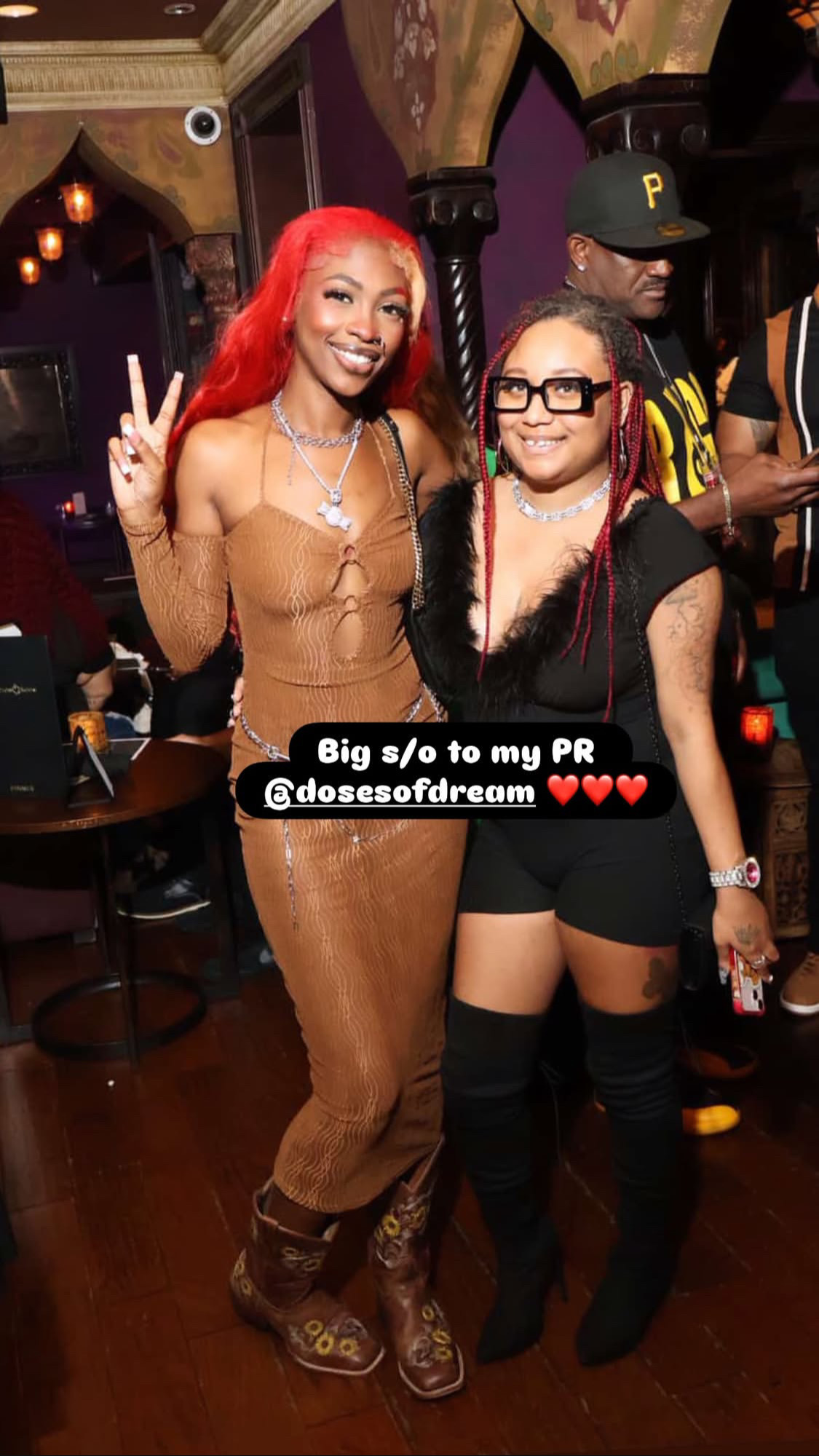Two women pose together in a lively indoor setting, one with red hair making a peace sign, the other with glasses smiling.