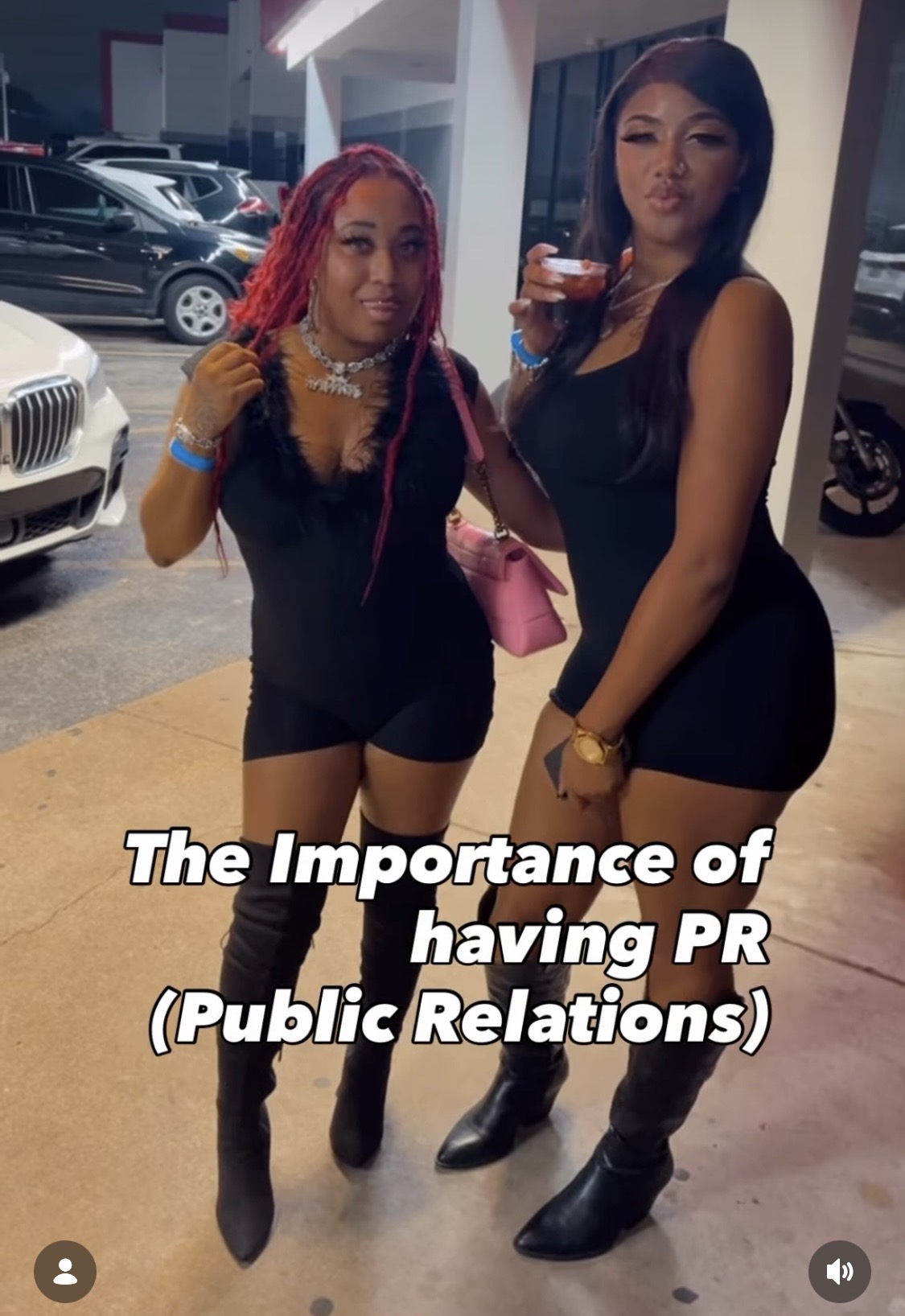 Two women standing indoors, one with pink braided hair and the other with long dark hair, holding drinks.