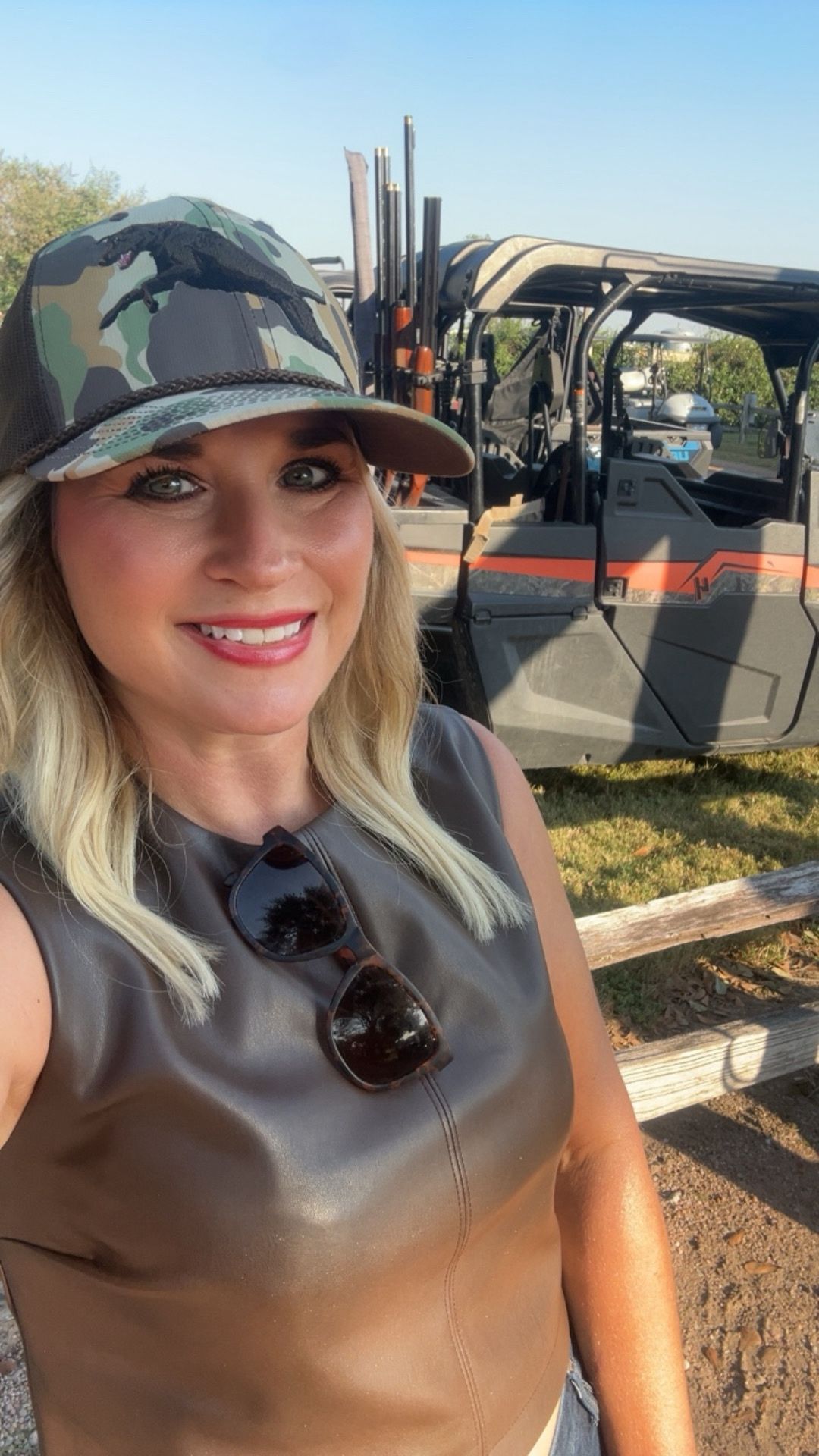 Woman outdoors wearing camouflage hat and sunglasses, smiling, with a vehicle and equipment in background.