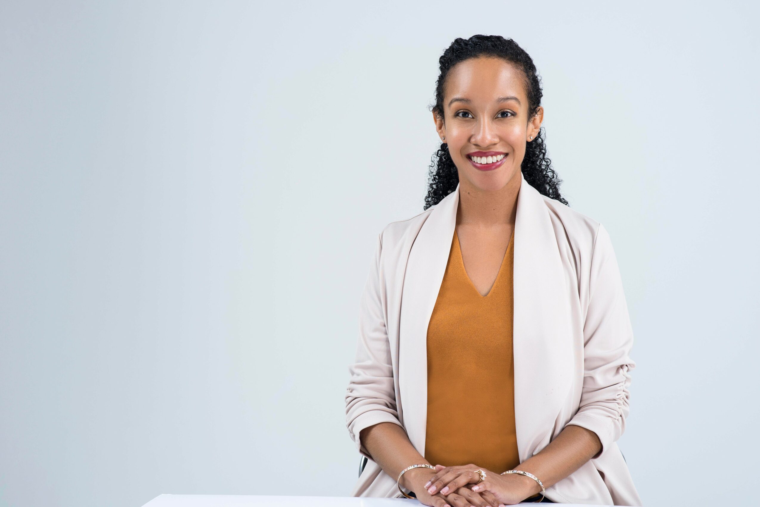 Smiling woman with curly hair, wearing a beige blazer and mustard top, standing against a plain light background.