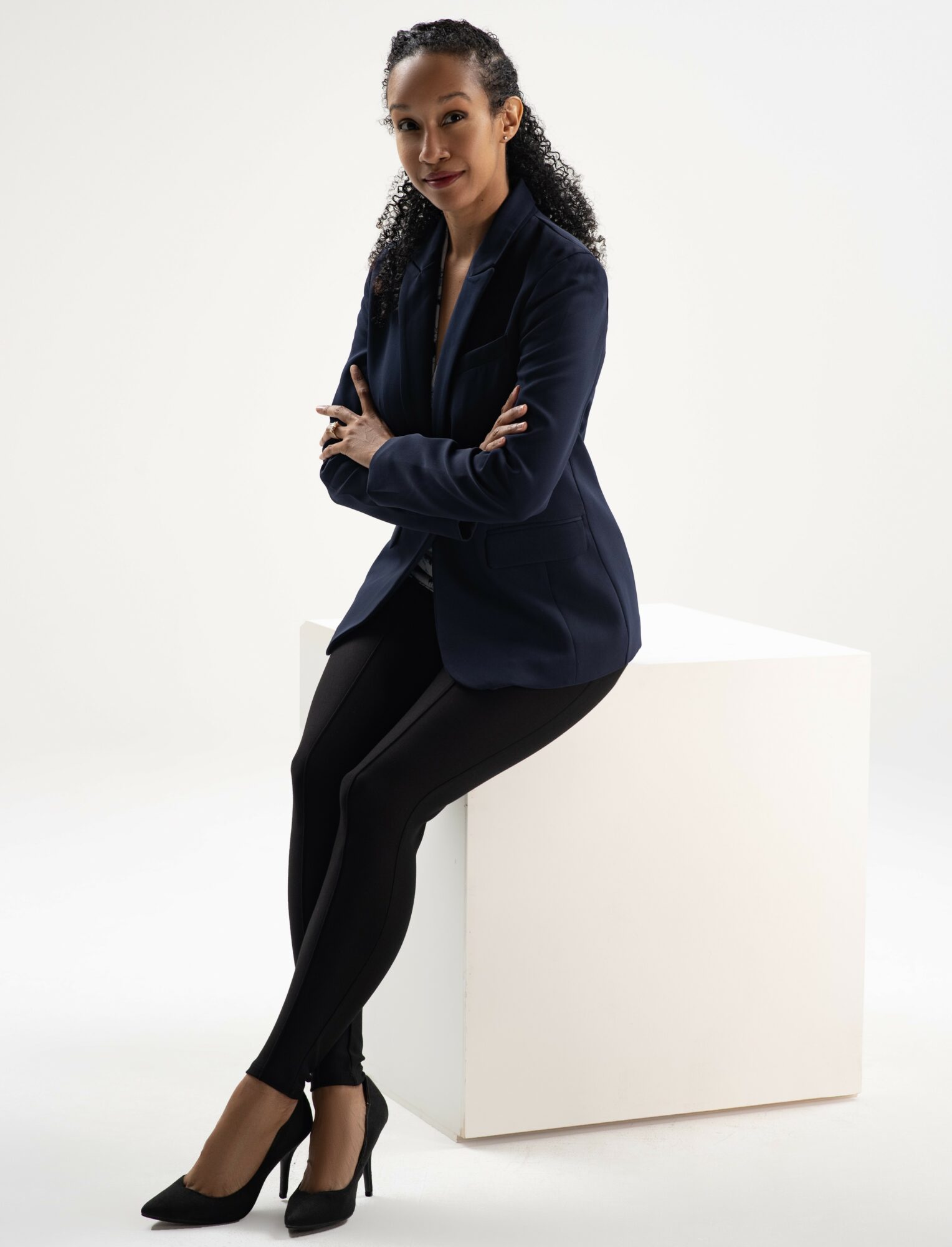 Woman sitting on a white block, wearing a dark blazer, black pants, and high heels, with arms crossed and smiling.