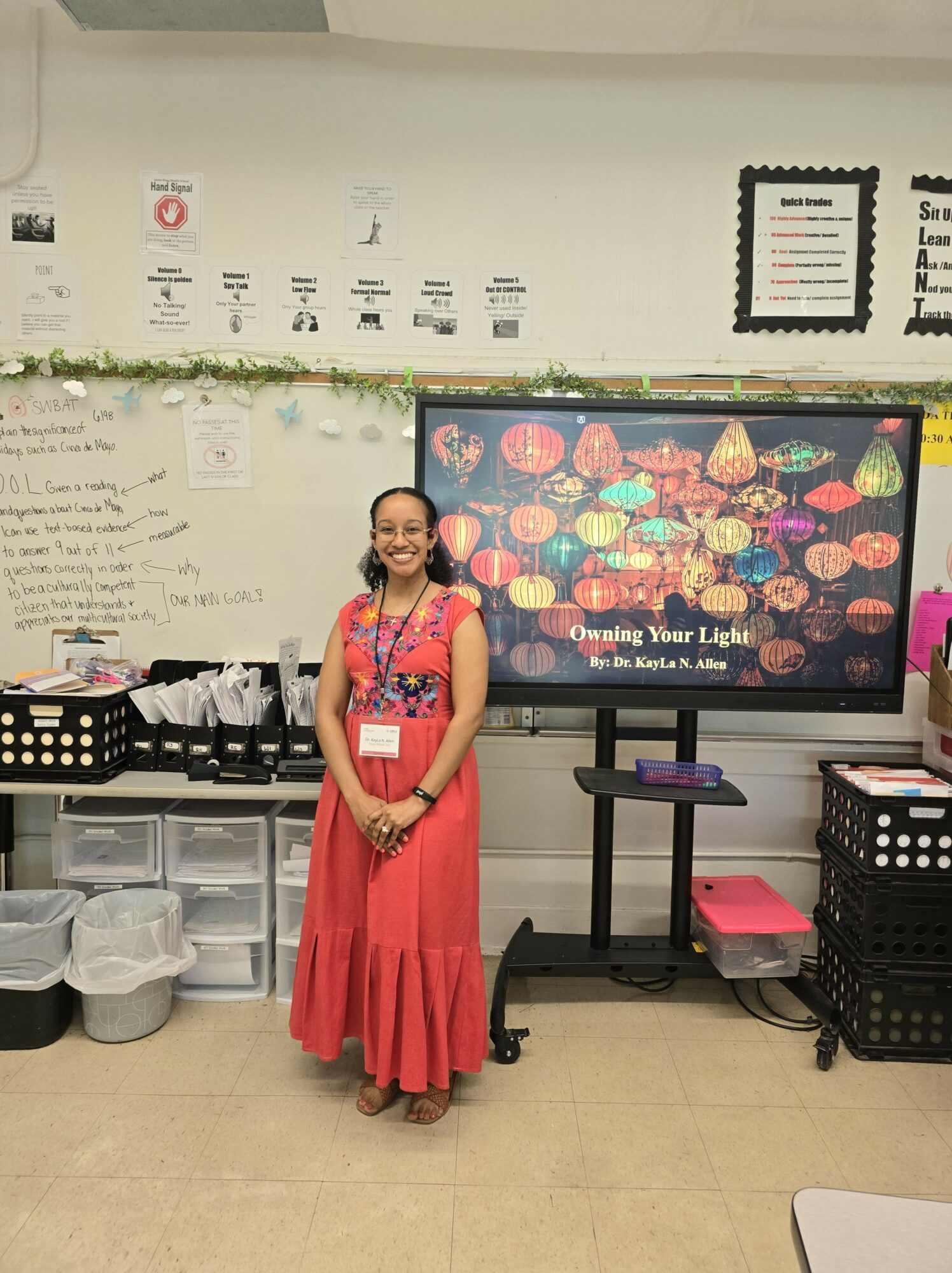 Smiling woman in a pink dress and floral top standing in classroom with whiteboard and digital screen behind her.