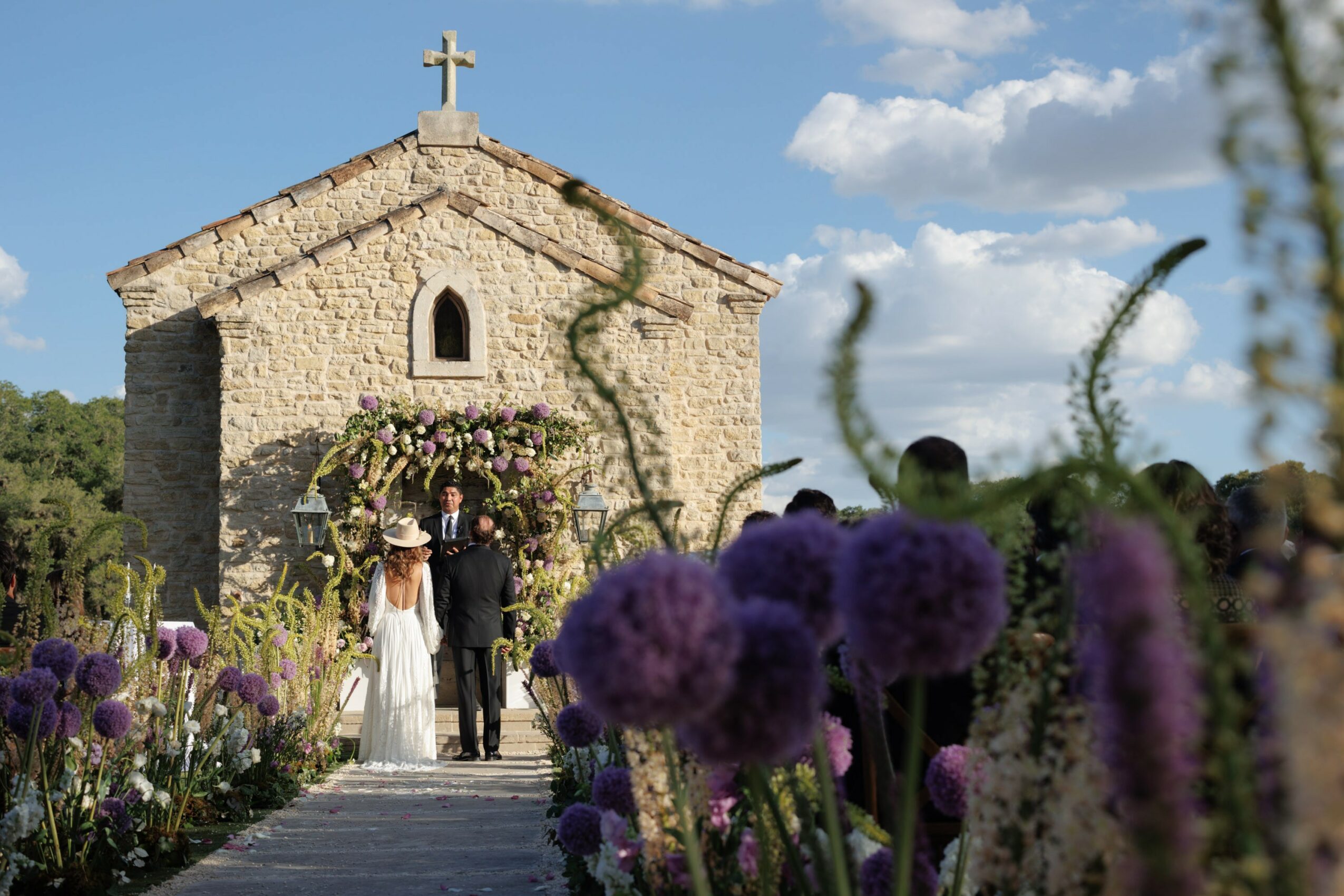Wedding ceremony outside a stone church with a cross, surrounded by flowers and a blue sky with clouds.