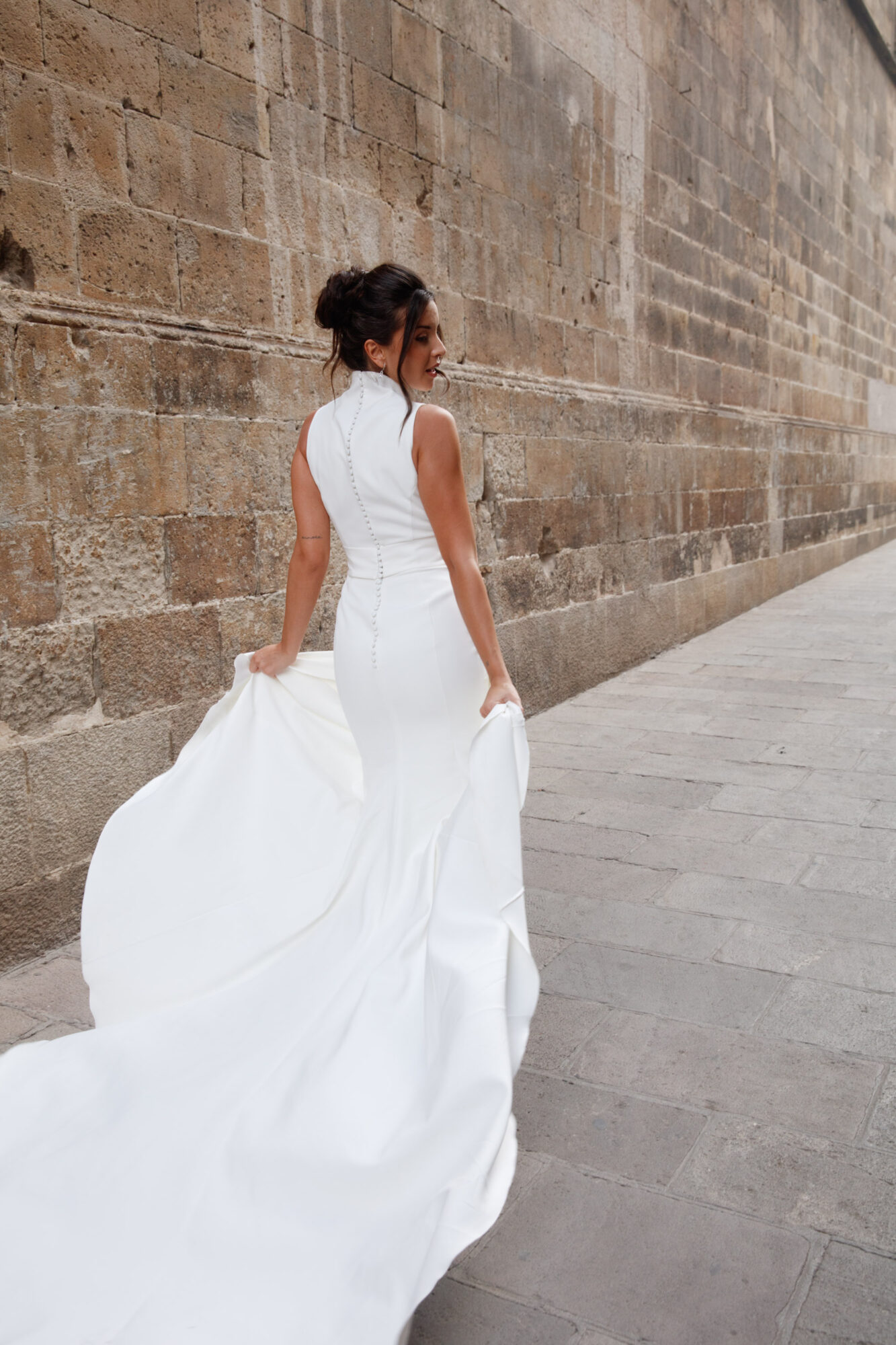 Woman in a white dress walking along a stone wall, holding the train of her dress.