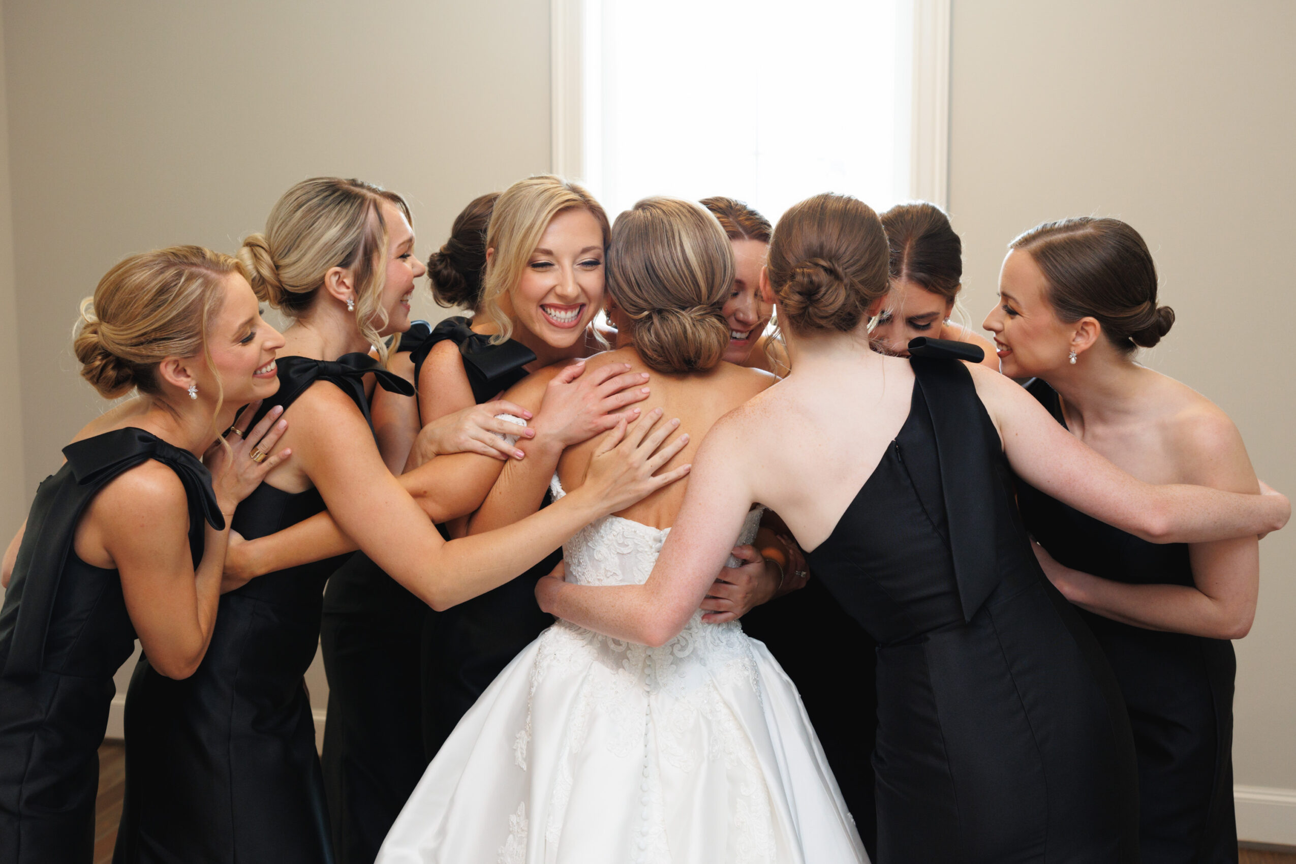 Group of women in black dresses hugging and smiling, celebrating in a room with light-colored walls and window.