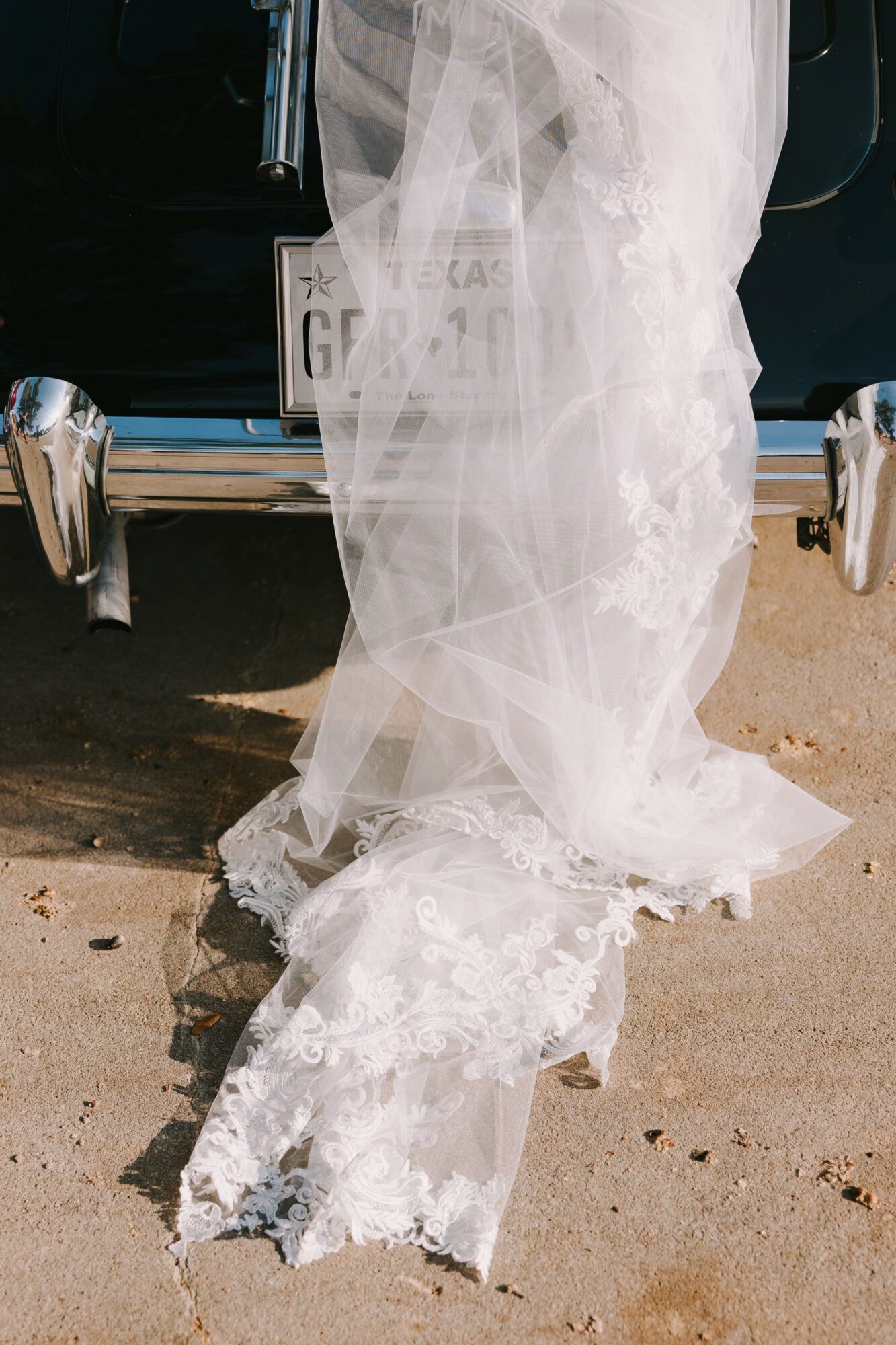 White lace and tulle fabric draped over a vehicle's bumper, partially covering a license plate, on a sandy surface.