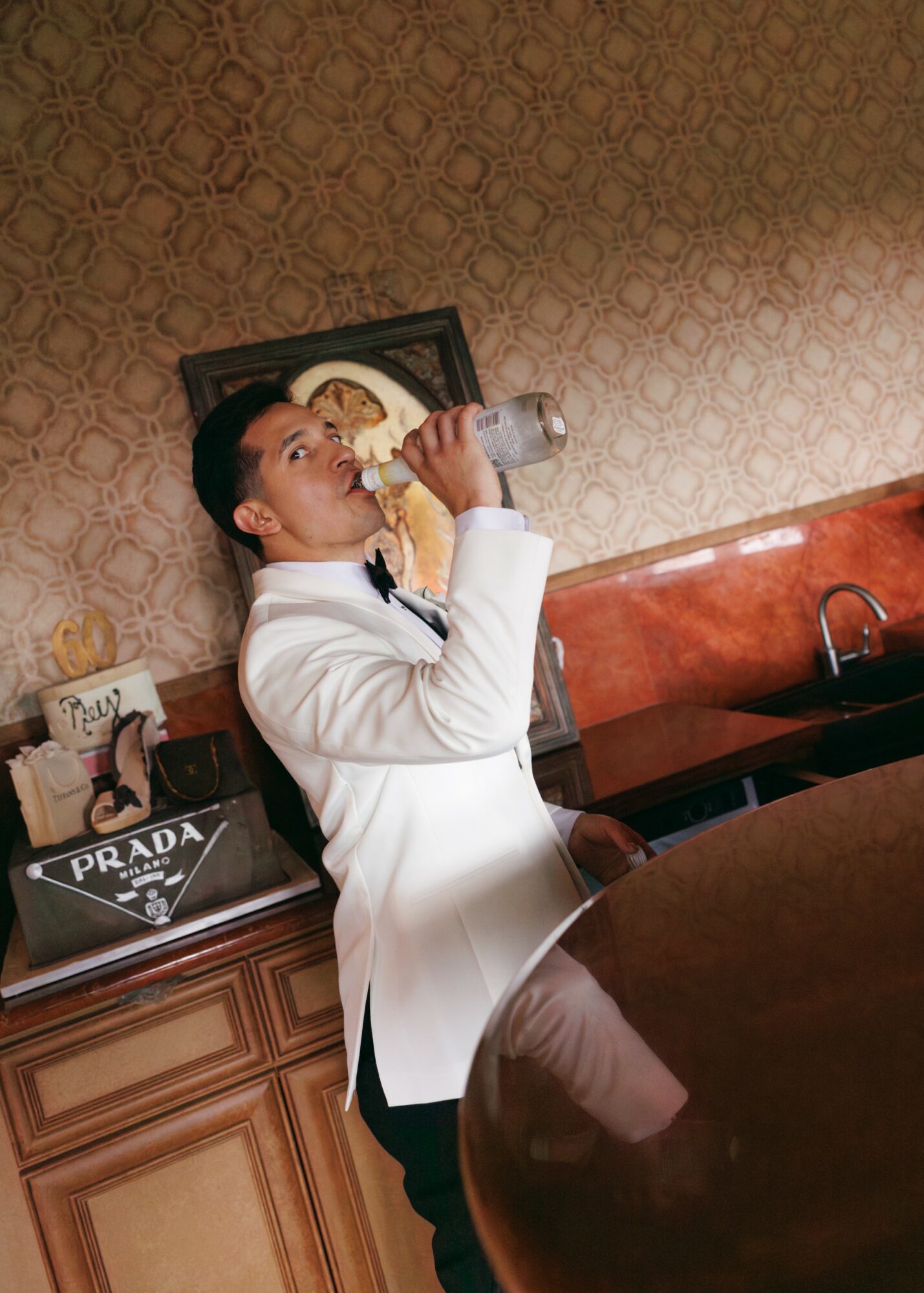 Person in white coat drinking from a bottle indoors, with a patterned wall and wooden furniture in background.