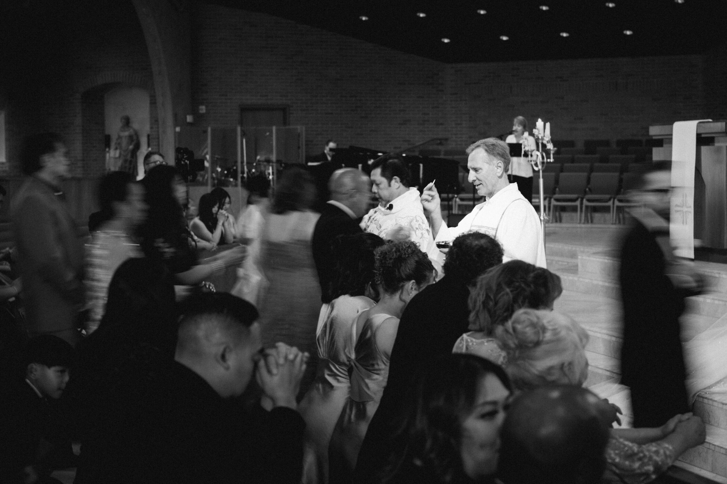 People gathered in a large indoor space, some standing and others sitting, with a stage and musical instruments in the background.