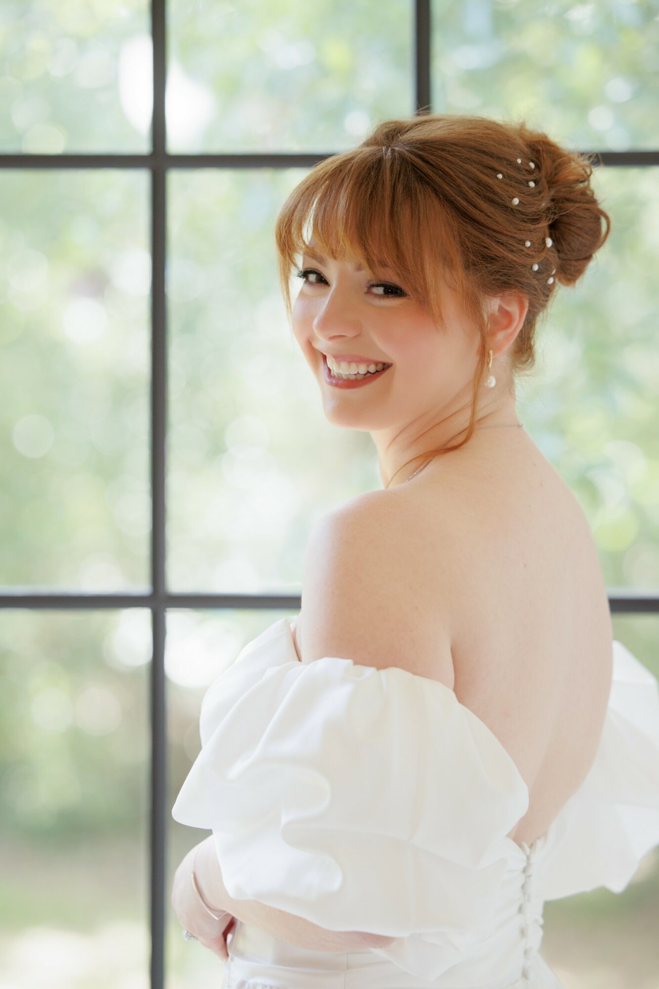 Young woman with red hair styled in an updo, smiling, wearing an off-shoulder white dress, standing near a window.
