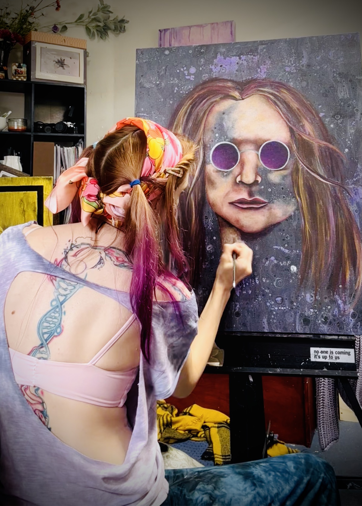 Person with pink and purple hair and glasses painting a large portrait of a woman with sunglasses.