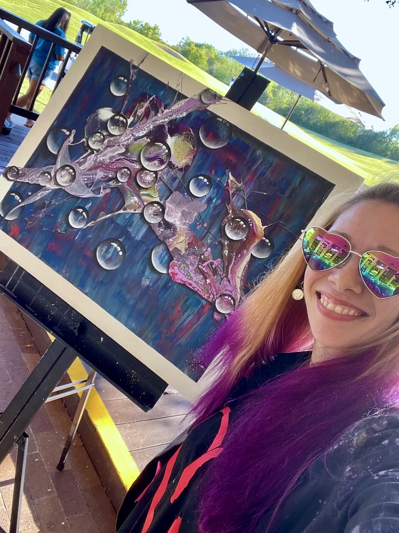 Person with colorful sunglasses smiling next to an abstract painting with bubbles and vibrant colors outdoors.