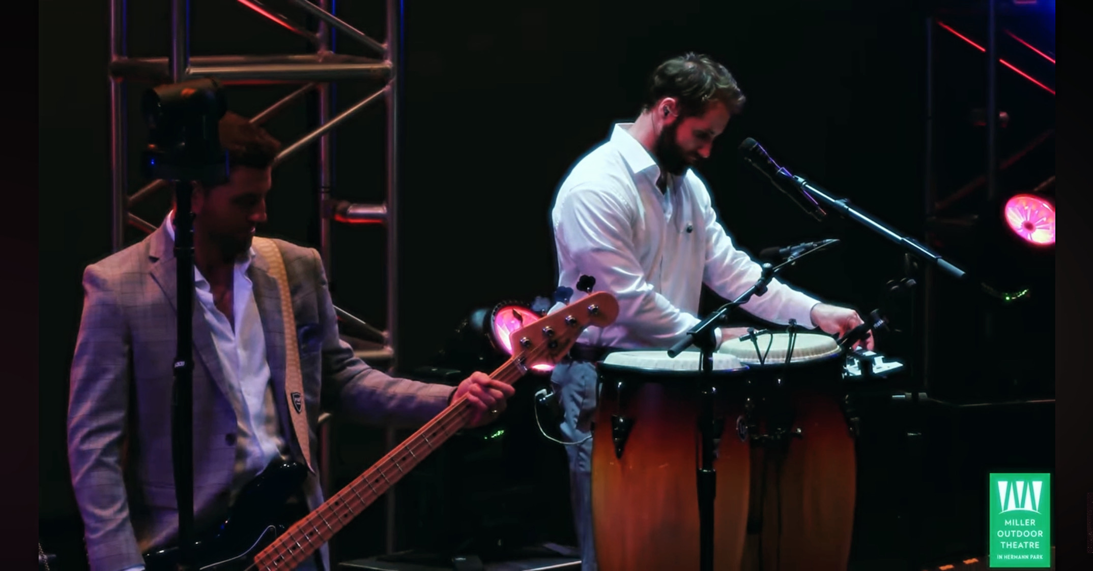 Two musicians perform on stage, one playing a bass guitar and the other playing drums, with stage lighting in background.
