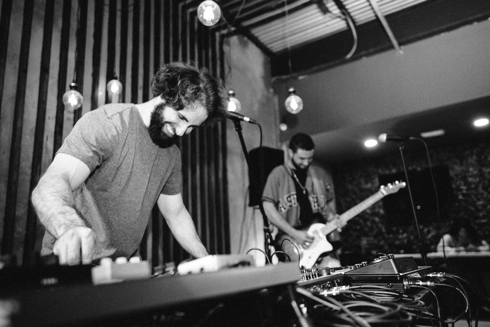 Two musicians perform on stage with equipment, one playing keyboard and the other guitar, under hanging lights.