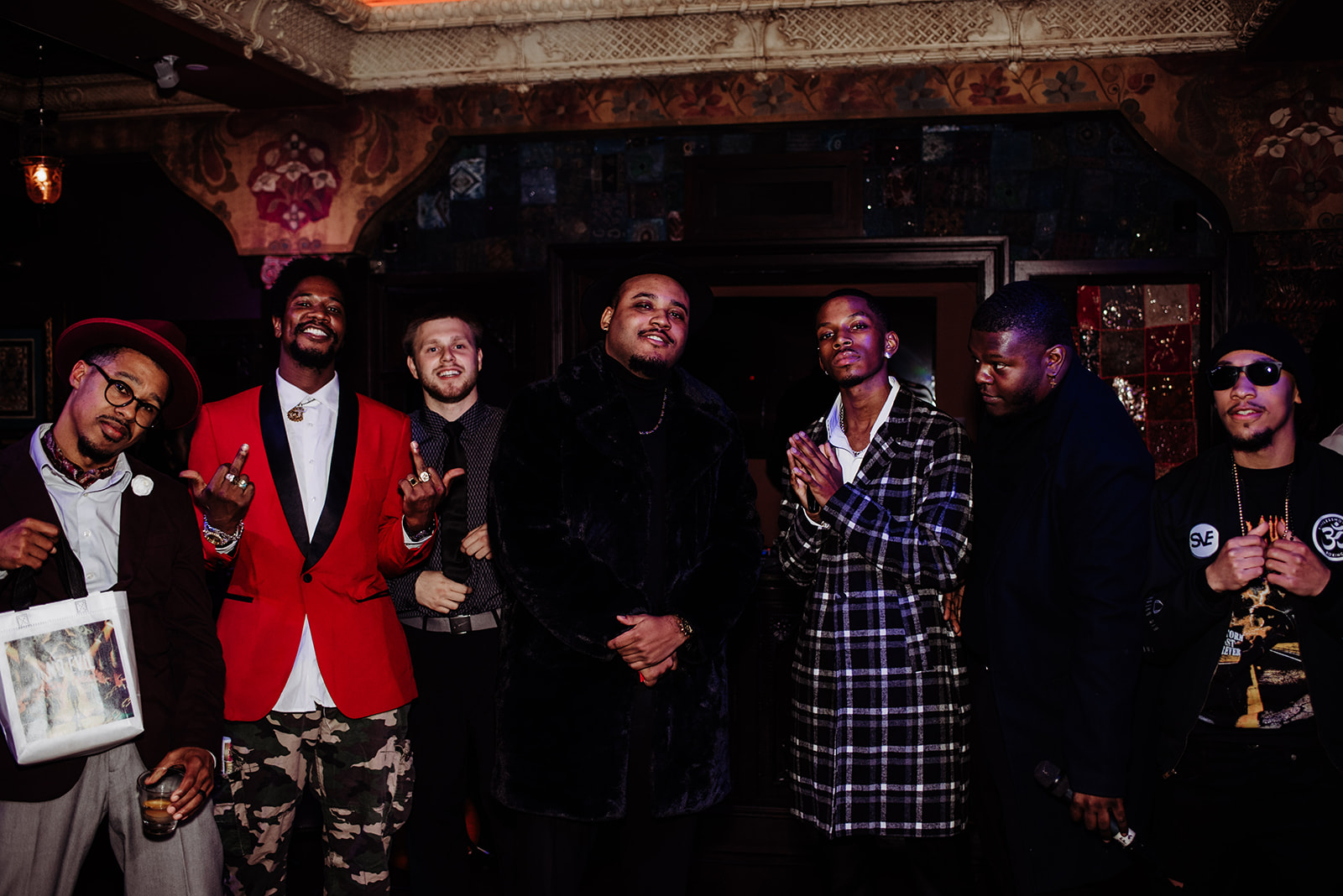 Group of seven people in a dimly lit room, some holding drinks, dressed in stylish clothing, smiling and posing for the photo.
