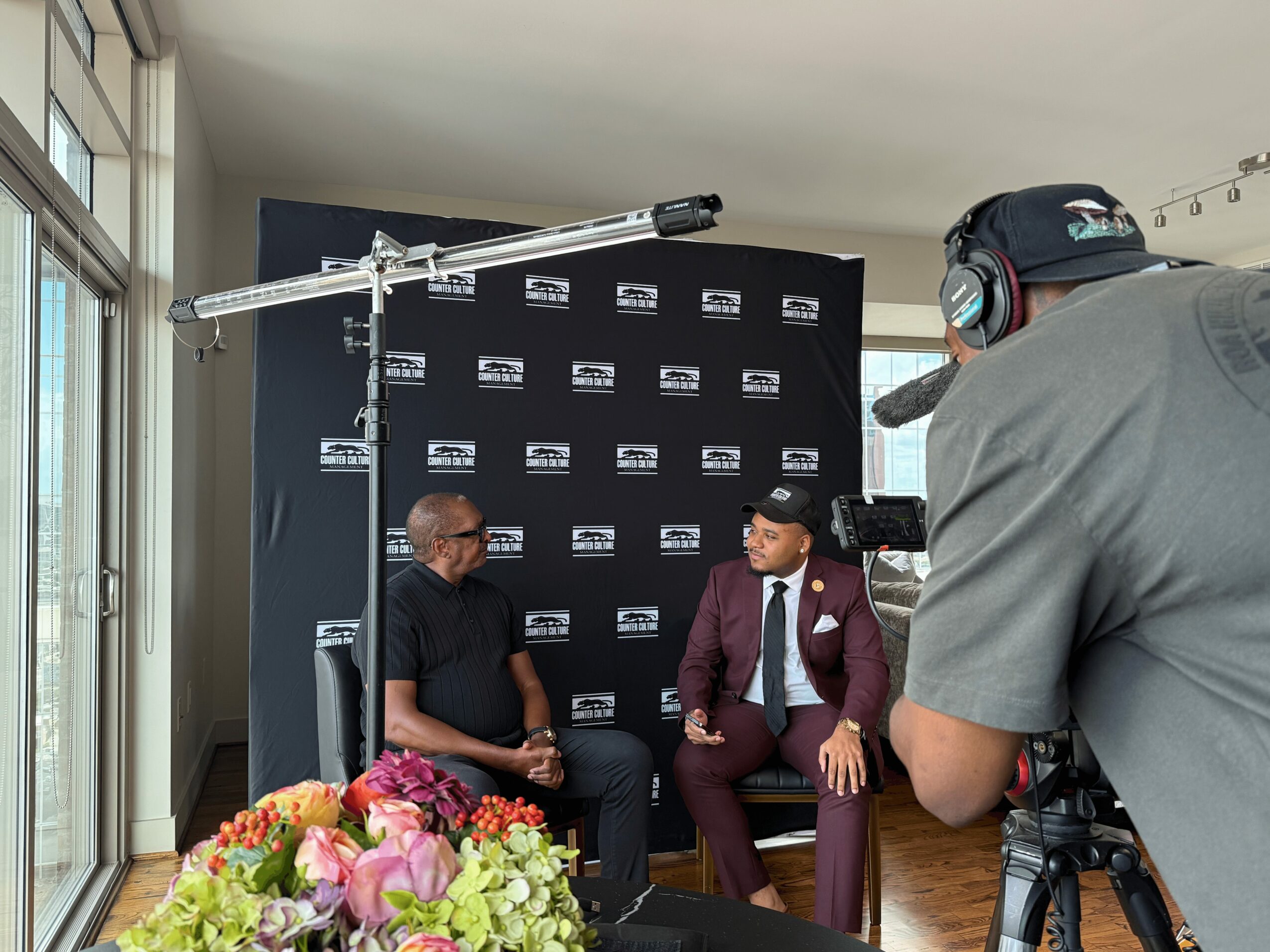 Two men seated in front of a black backdrop with logos, one in casual clothing and the other in a suit, during an interview.