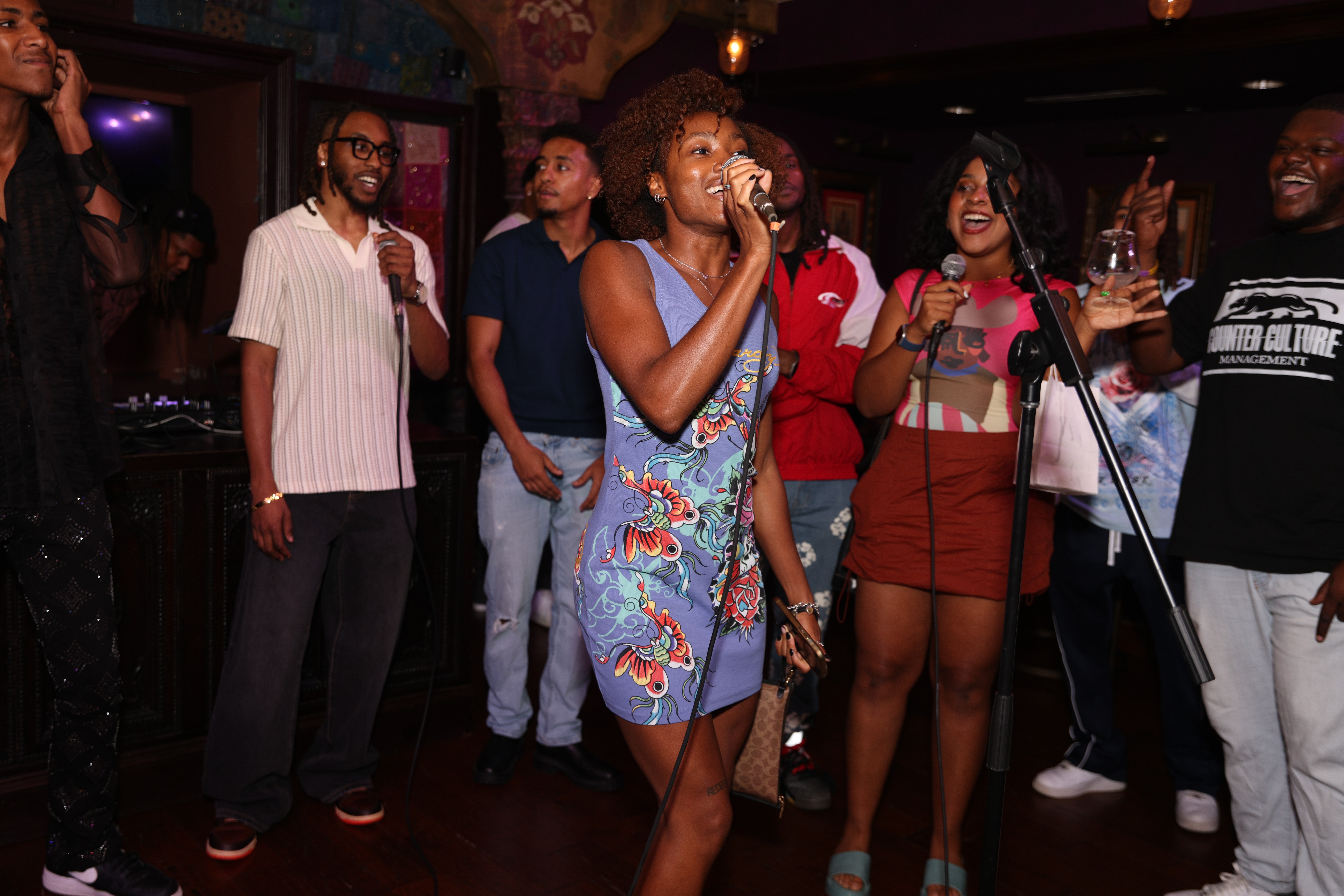 Group of people at a party, some singing and holding drinks, in a dimly lit room with dark walls.