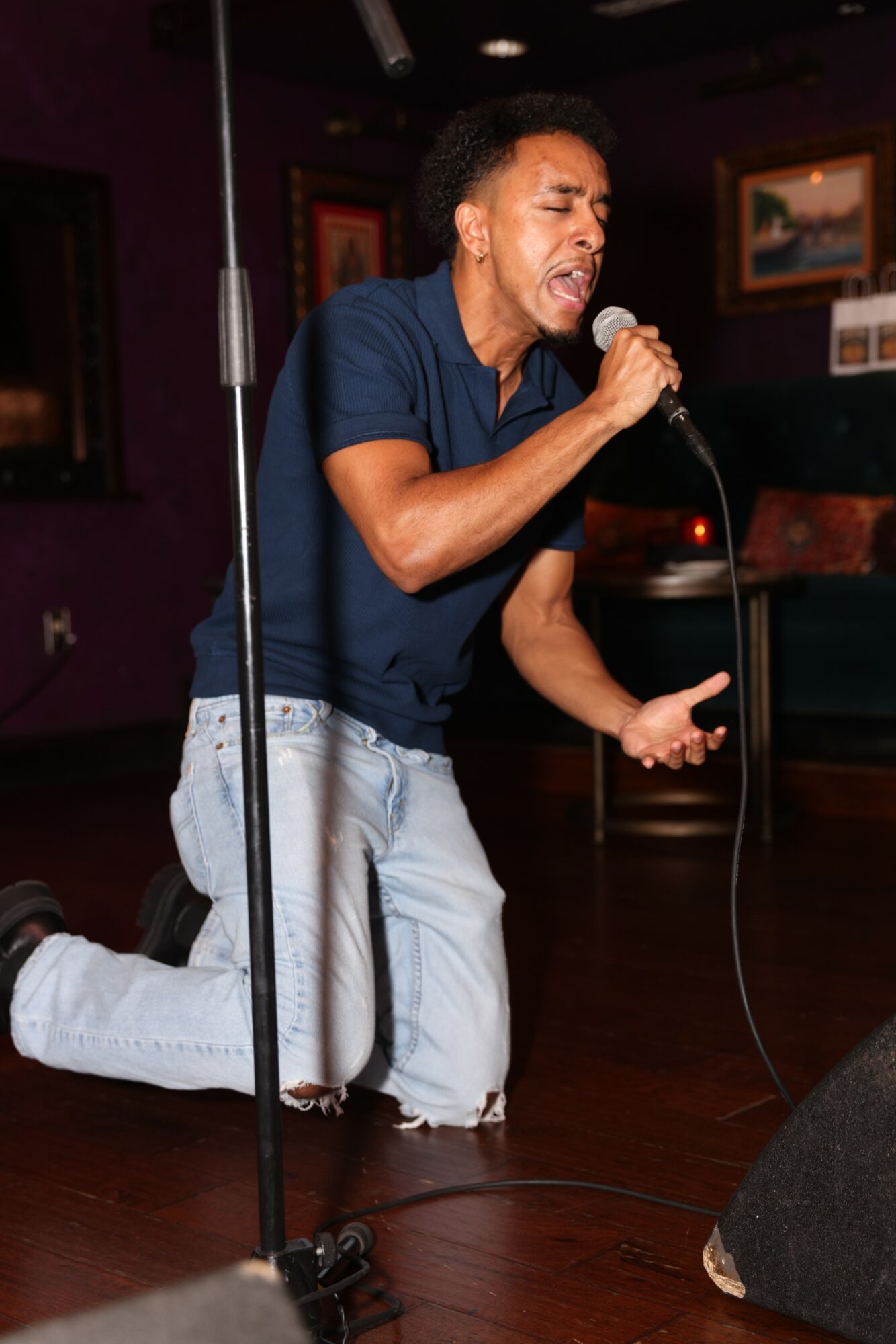 Person kneeling on stage, singing into microphone with eyes closed, in a room with framed pictures on wall.