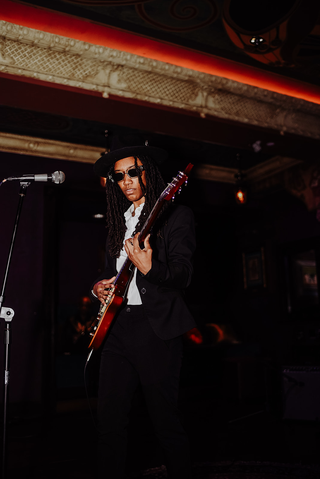 Person with long dreadlocks wearing sunglasses and a suit, holding an electric guitar on stage.